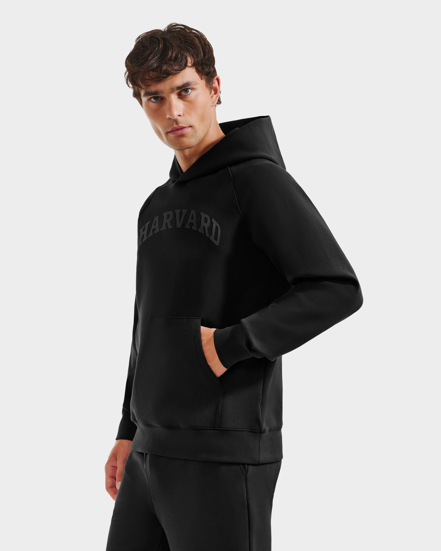A young man wears the Rudsak BRIAR R HV - H525506 Black hoodie, standing with one hand in his pocket and gazing to the side against a plain, light background.