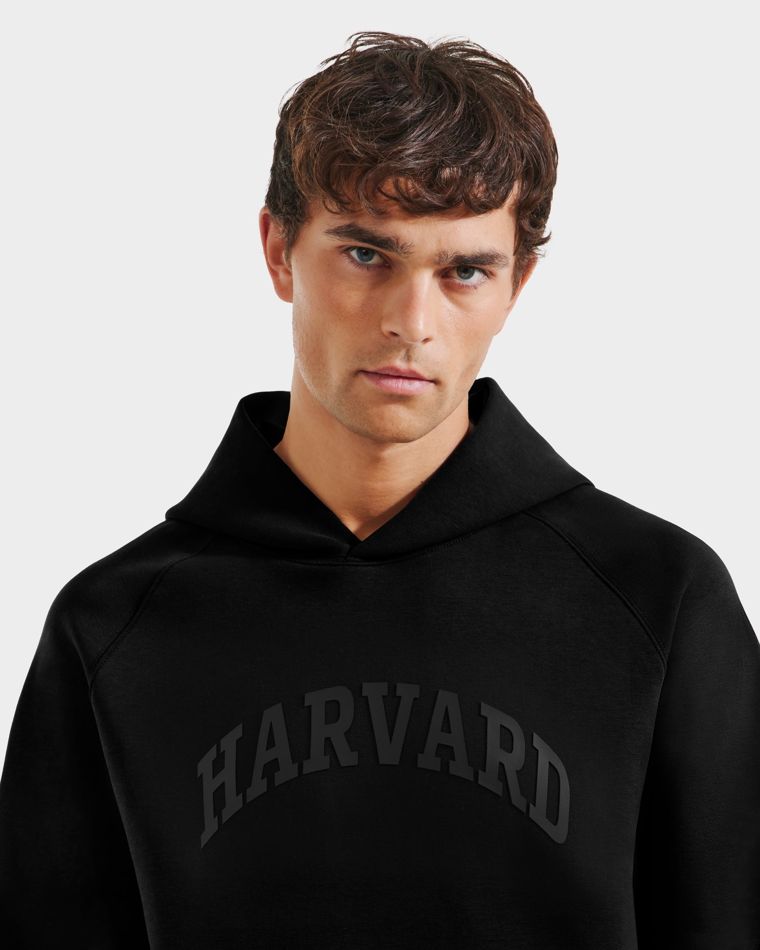 A young man with short brown hair wears a black Rudsak BRIAR R HV - H525506 hoodie, bold HARVARD letters across his chest, as he stands against a plain light background.