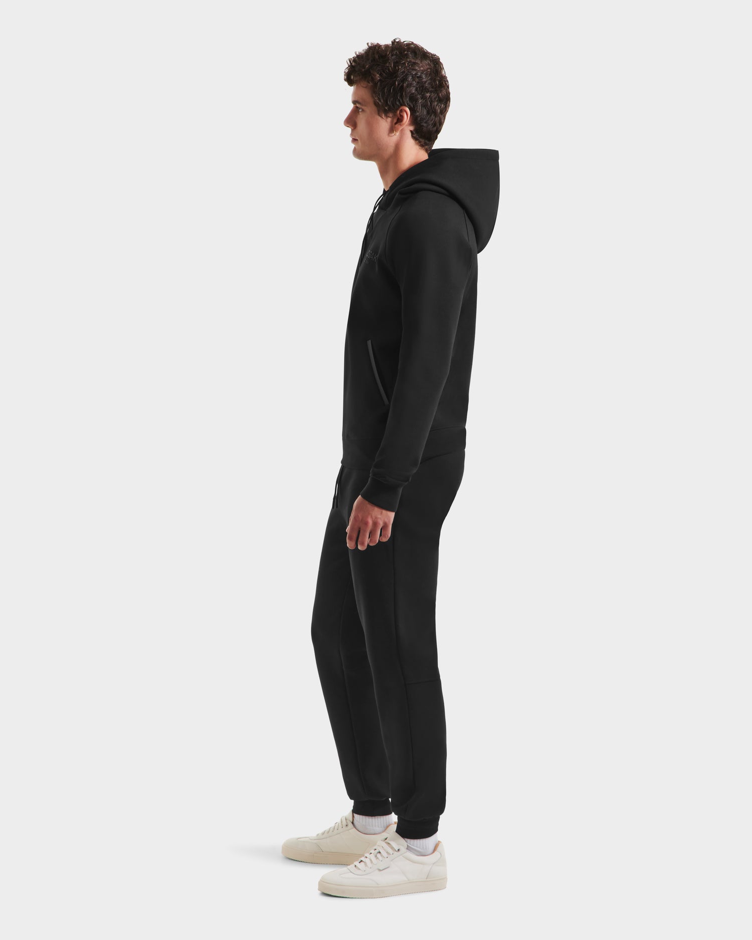 A man in profile wears the Rudsak BROOKLYN - 6525593 Black hoodie, styled with black jogger pants and white sneakers, embodying effortless urban style against a plain white background.