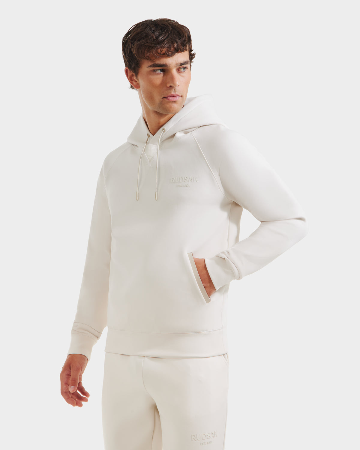 A man models the Rudsak BROOKLYN - 6525593 Vanilla hoodie and sweatpants set, standing against a white background with one hand in his pocket, embodying a clean urban streetwear look as he gazes to the side.