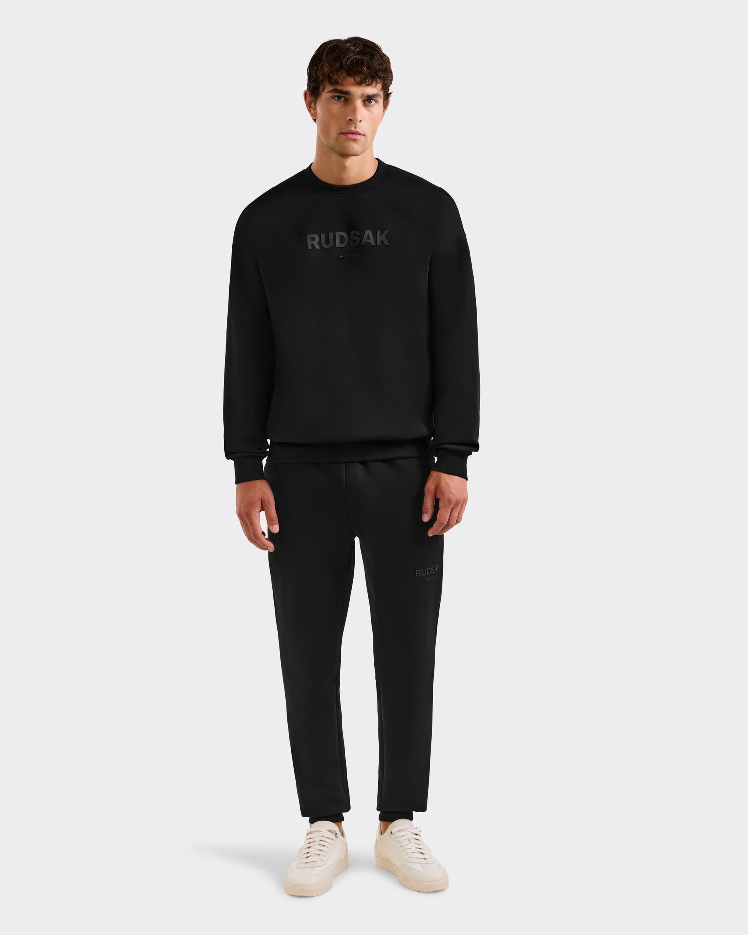 A man faces forward in a black Rudsak BRYN - 9524007 crewneck sweatshirt and matching sweatpants, paired with white sneakers against a plain light gray background.
