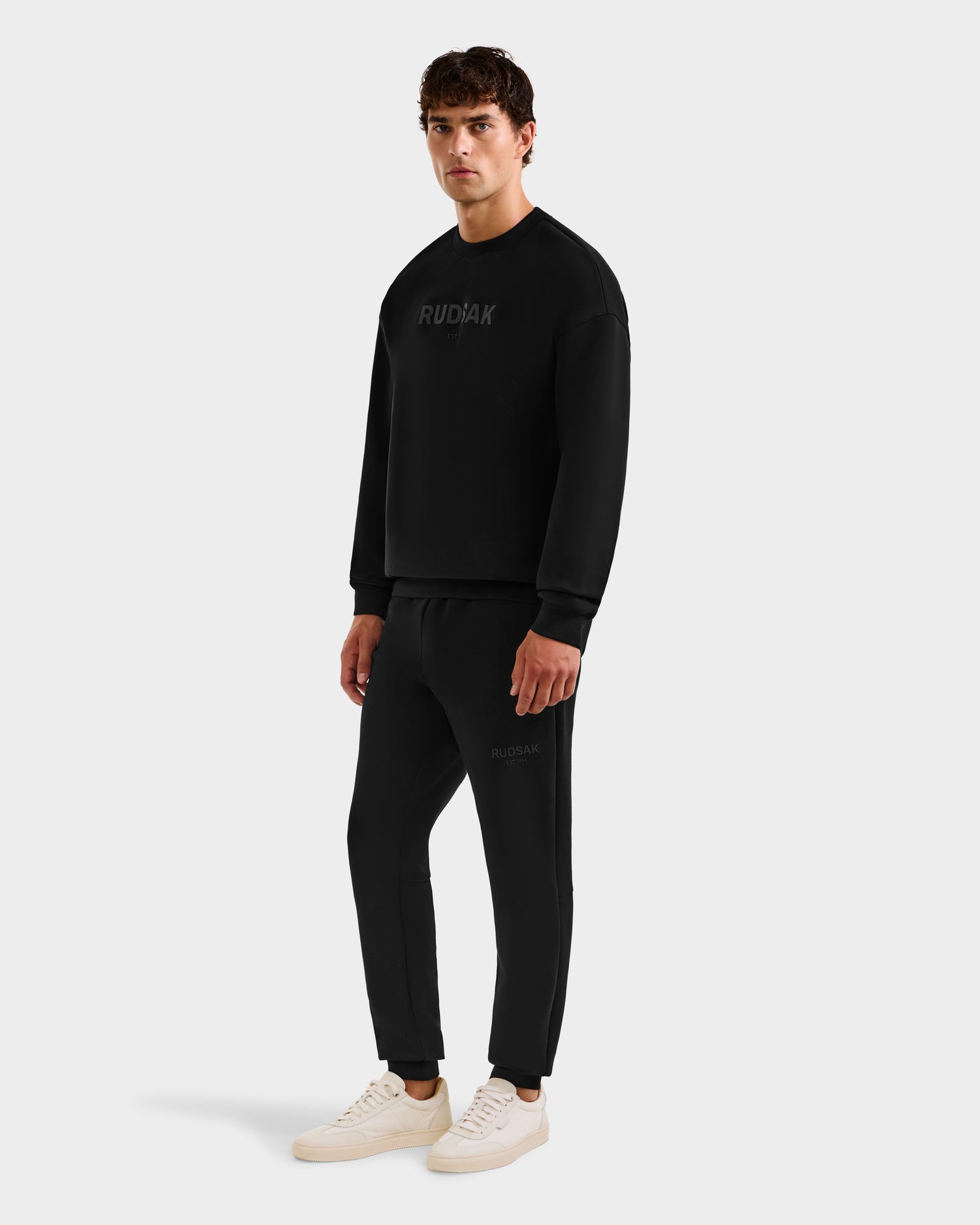 A man wears the Rudsak BRYN - 9524007 Black M crewneck sweatshirt and joggers in genderless sizing, styled with white sneakers. Subtle branding adds a finishing touch to his everyday look against a plain light background.