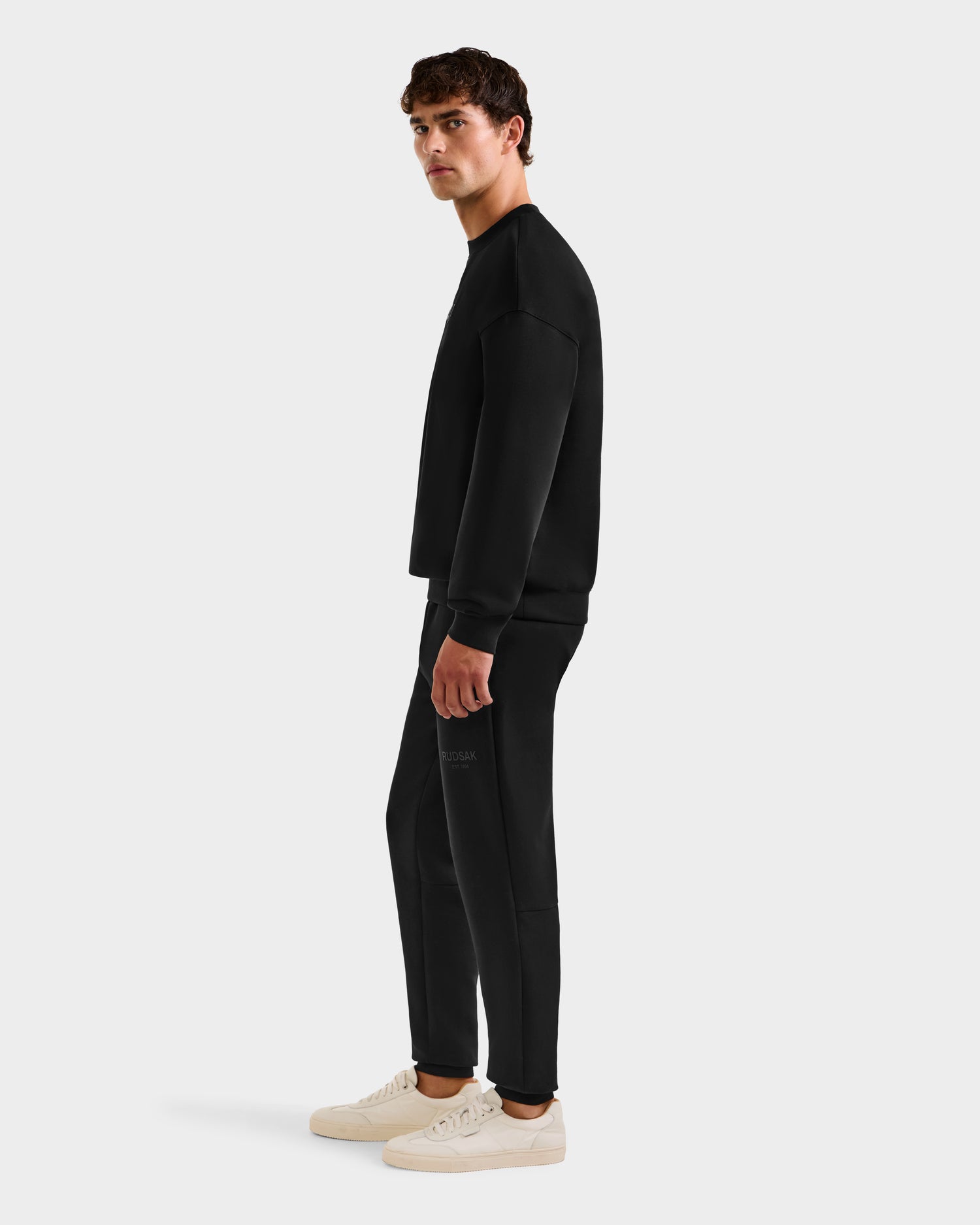 A man wears the Rudsak BRYN - 9524007 Black M cotton crewneck sweatshirt and matching sweatpants with white sneakers, posing sideways against a plain light background. The outfit offers an everyday fit and genderless sizing.