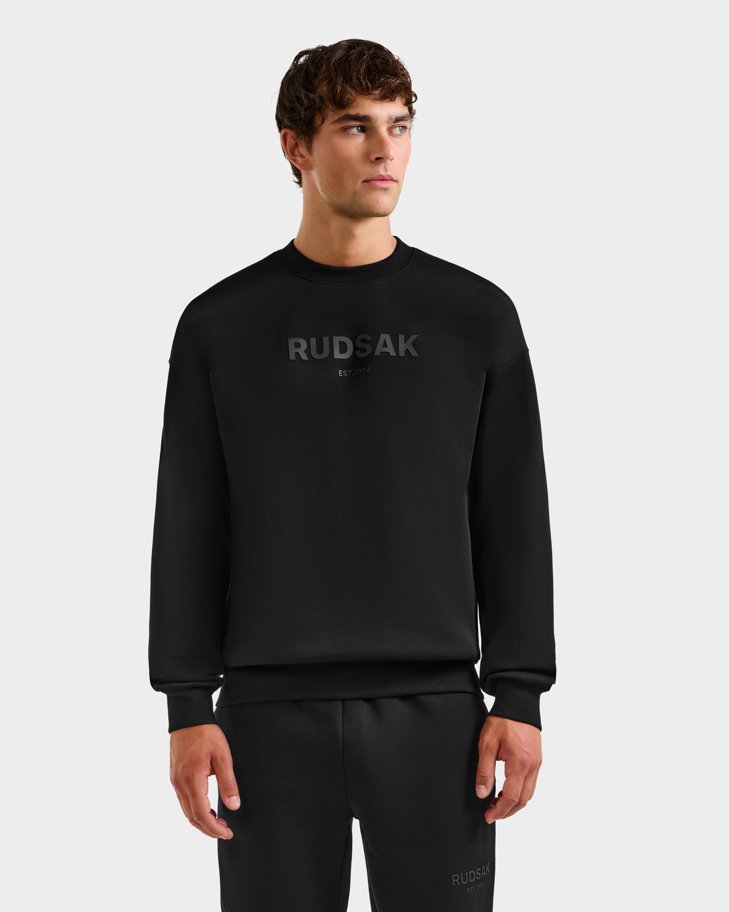 A young man in the Rudsak BRYN - 9524007 black cotton crewneck sweatshirt, featuring everyday fit and genderless sizing with "RUDSAK" on the chest, stands in black pants against a plain light gray background.