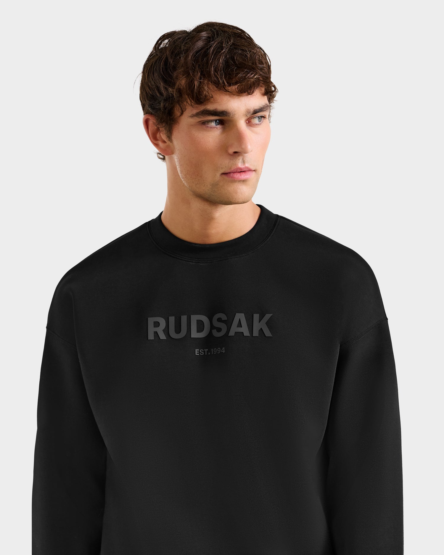 A young man with brown hair wears the Rudsak BRYN - 9524007 Black M cotton crewneck sweatshirt featuring RUDSAK EST. 1994 in dark lettering, styled in a relaxed fit as he looks slightly left against a plain light background.