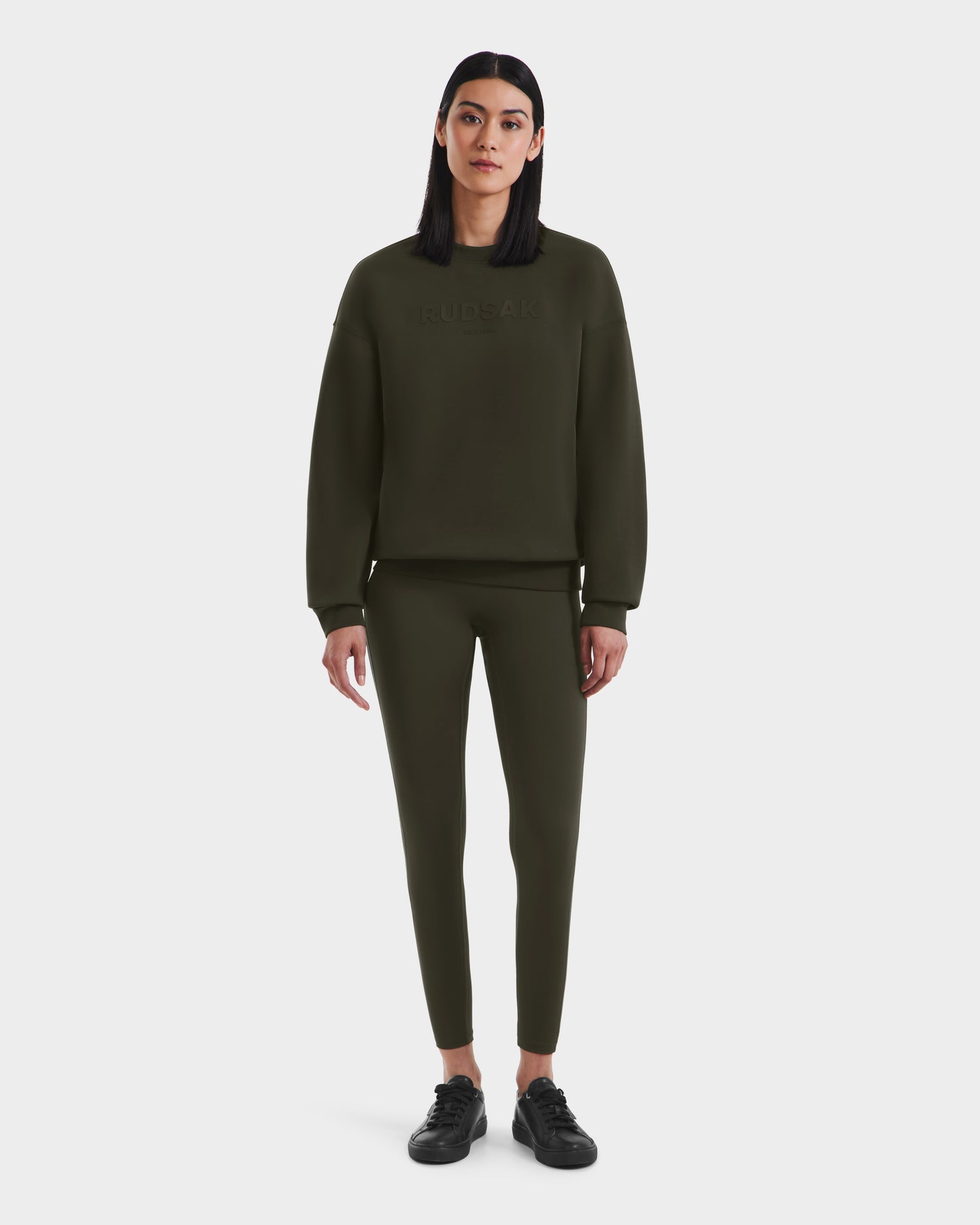 A woman with straight, dark hair faces forward wearing the Rudsak BRYN - 9524007 Dark Olive W cotton crewneck sweatshirt and matching leggings with black sneakers, set against a plain white background.