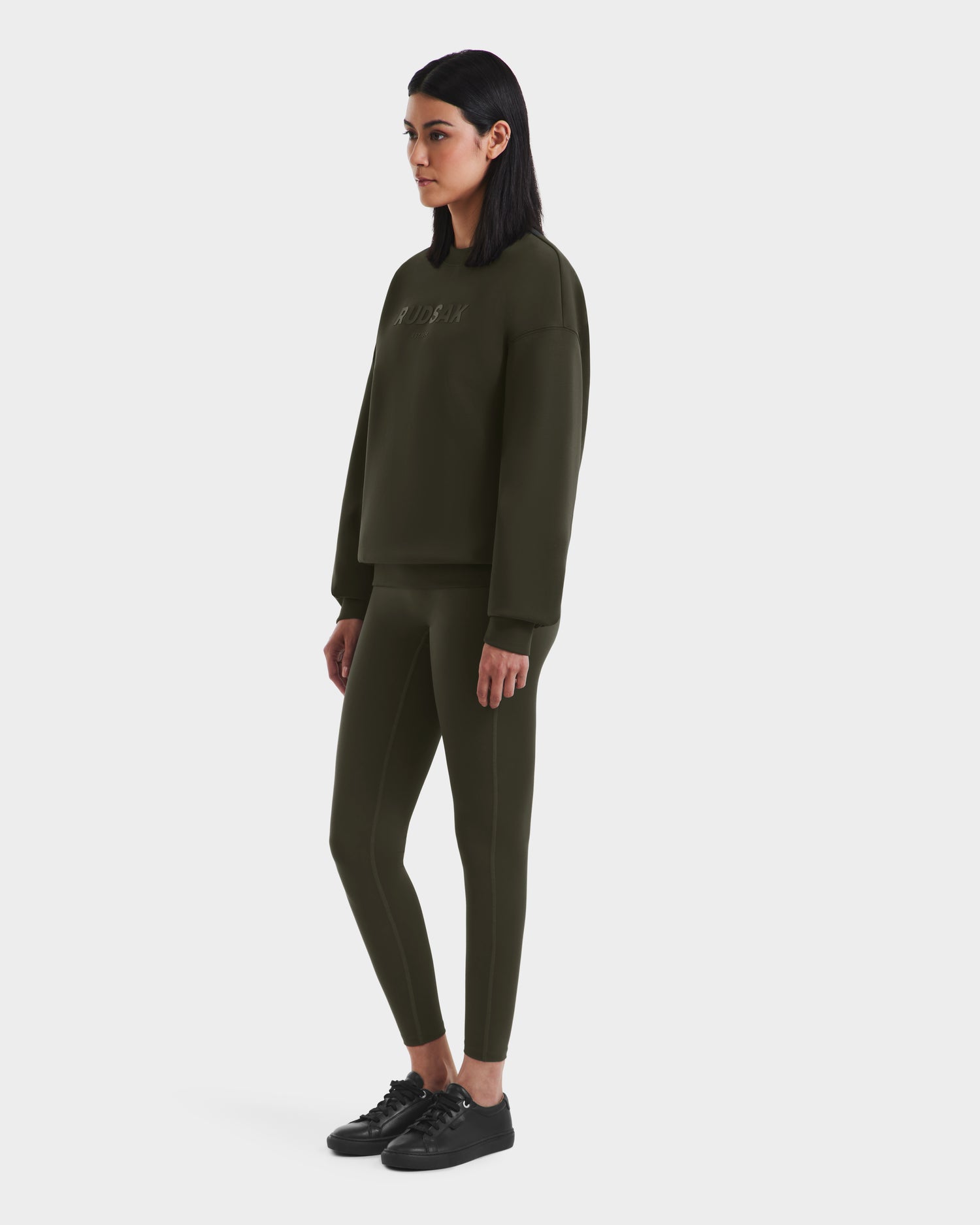 A woman with straight dark hair stands facing left, wearing the Rudsak BRYN - 9524007 Dark Olive W crewneck sweatshirt and matching leggings, paired with black sneakers, against a plain white background.