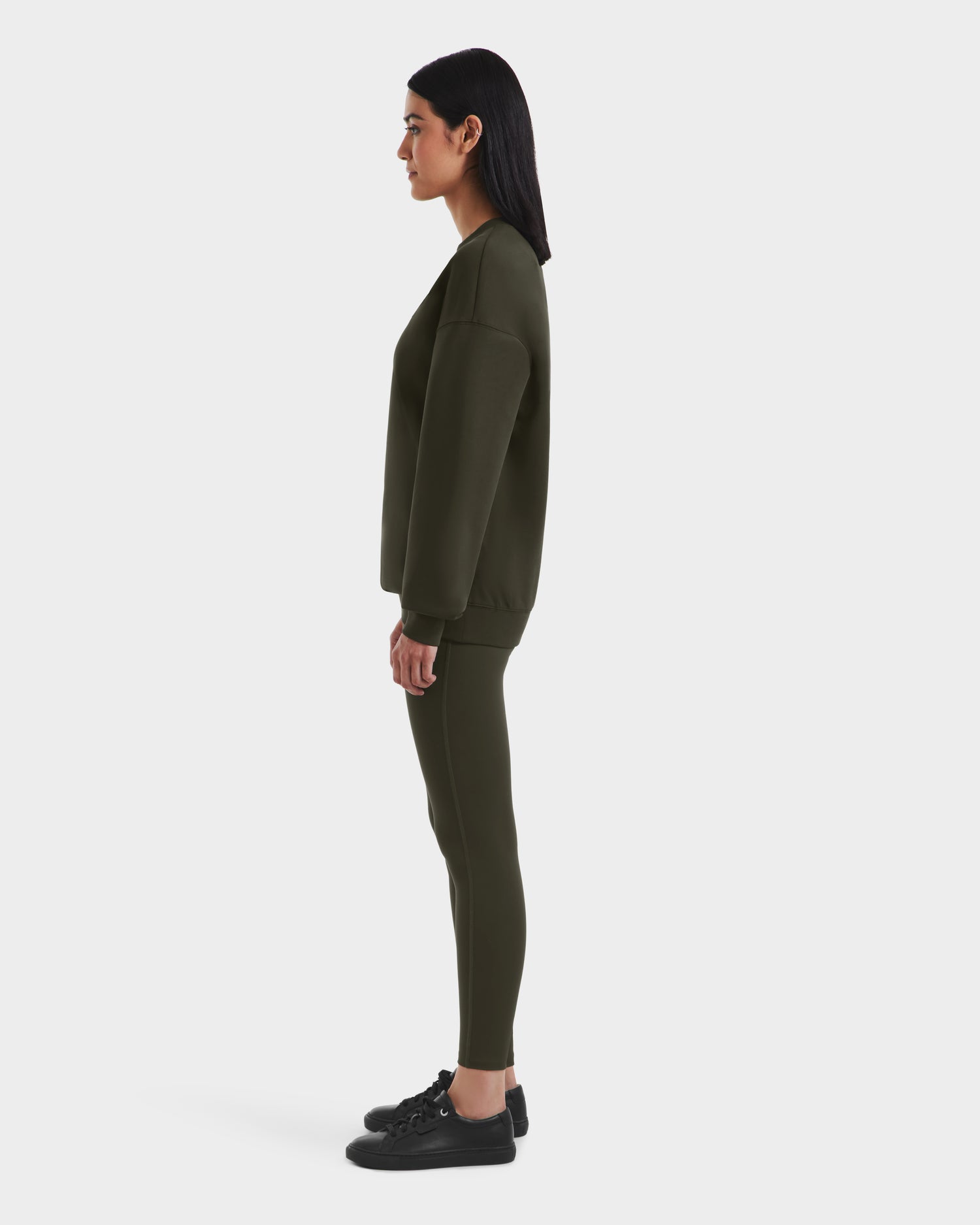 A woman in profile faces left, wearing the Rudsak BRYN - 9524007 Dark Olive W cotton crewneck sweatshirt with matching fitted pants and black sneakers, emphasizing its timeless style and genderless fit against a plain white background.