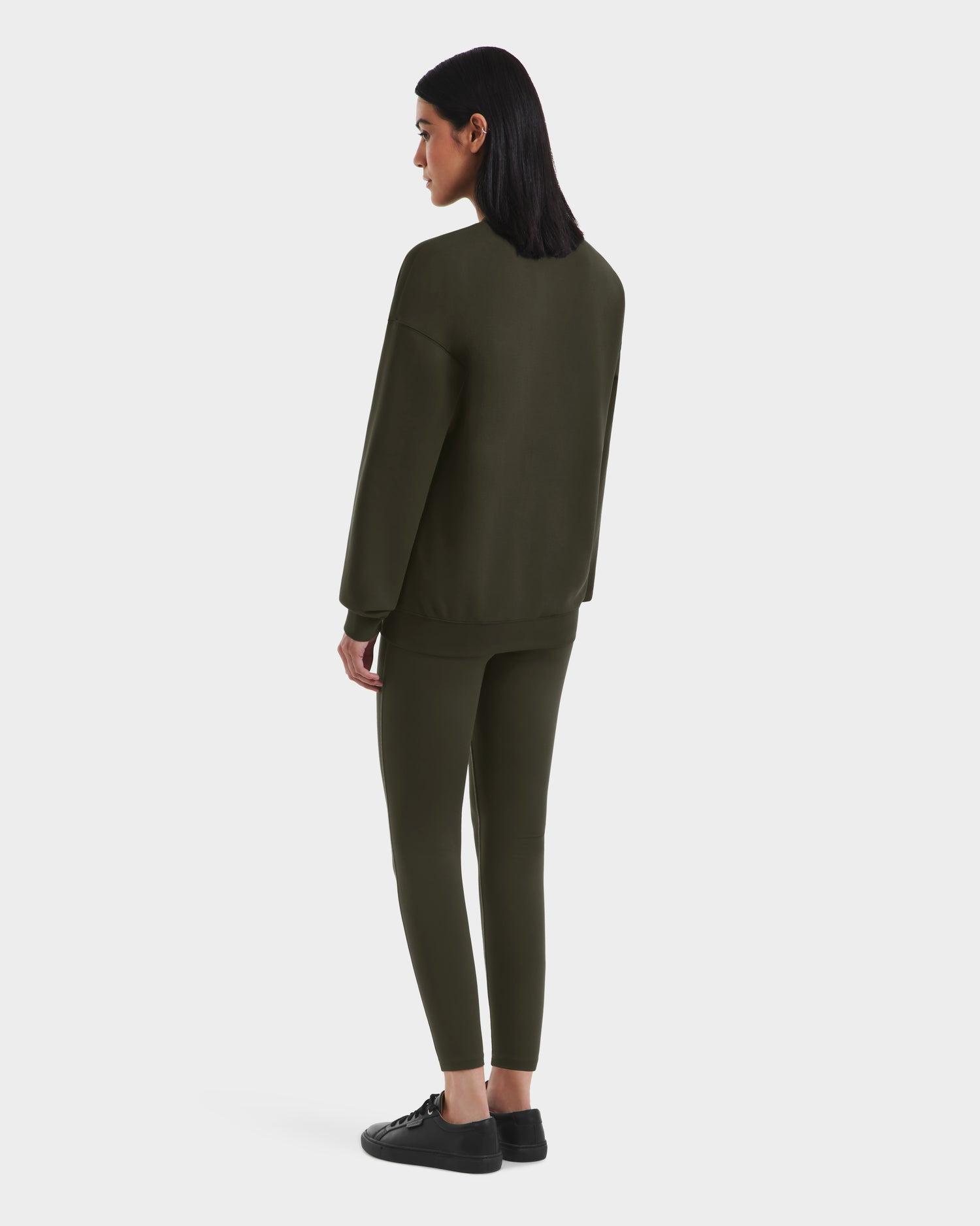 A person with shoulder-length dark hair faces away, wearing the Rudsak BRYN - 9524007 Dark Olive W cotton crewneck sweatshirt with matching leggings and black sneakers, set against a plain white background—a classic look for any style.