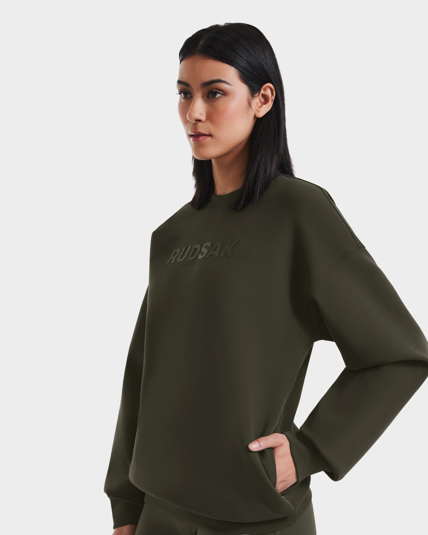 A woman with straight black hair models the Rudsak BRYN - 9524007 Dark Olive W, an oversized cotton crewneck sweatshirt with embossed branding, standing against a plain white background to highlight its classic style.