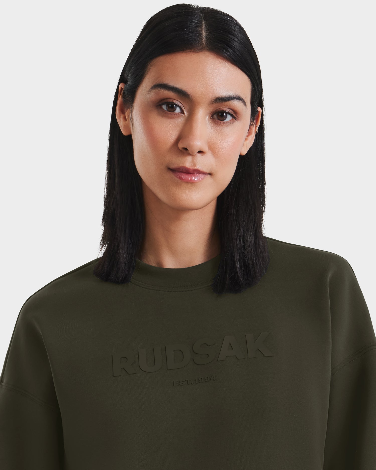 A woman with straight, shoulder-length black hair wears the Rudsak BRYN - 9524007 Dark Olive W crewneck sweatshirt and looks neutrally at the camera against a plain white background.