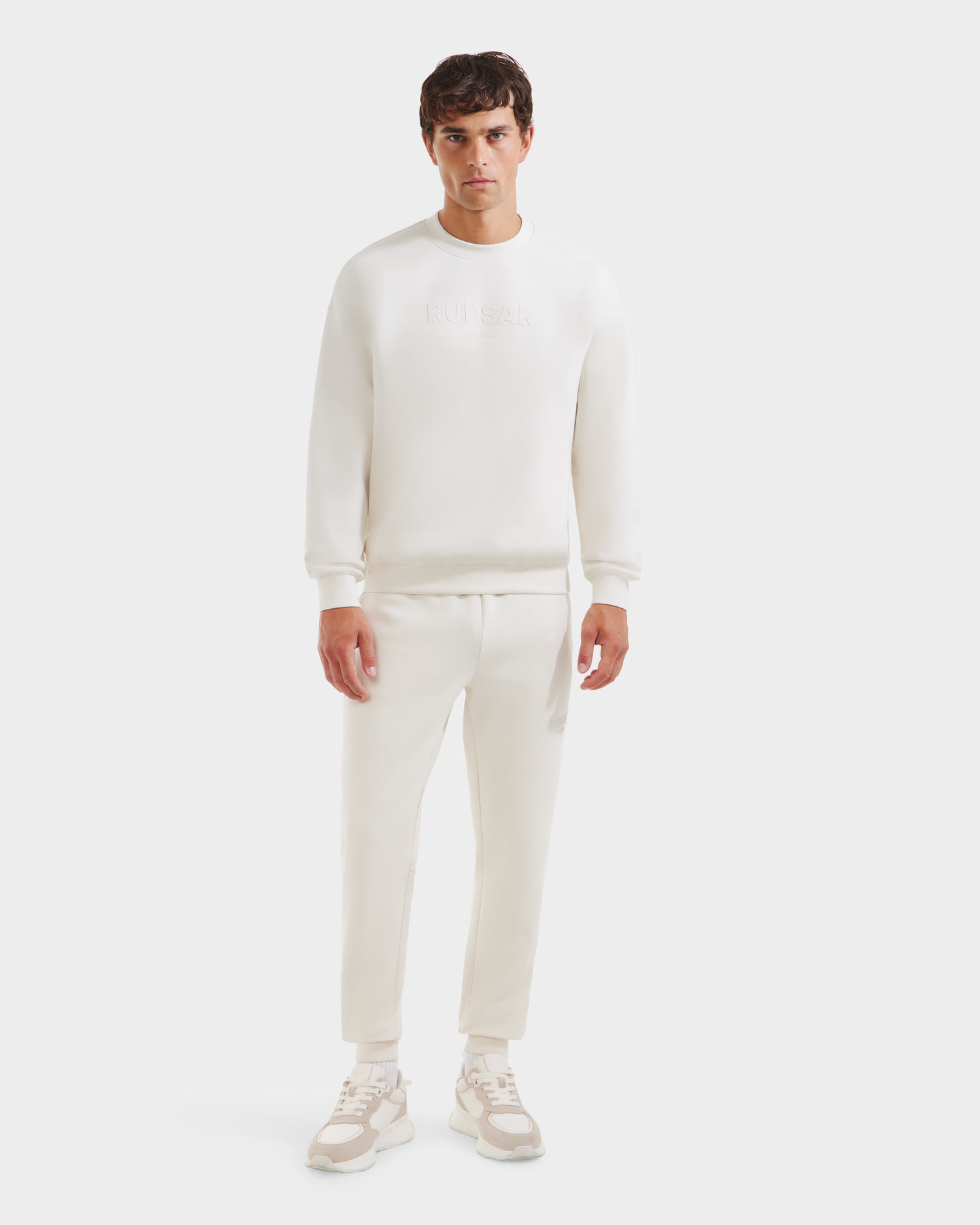 A man faces forward in a casual look, sporting Rudsak’s BRYN - 9524007 Vanilla M crewneck sweatshirt, jogger pants, and beige-and-white sneakers against a plain white background.