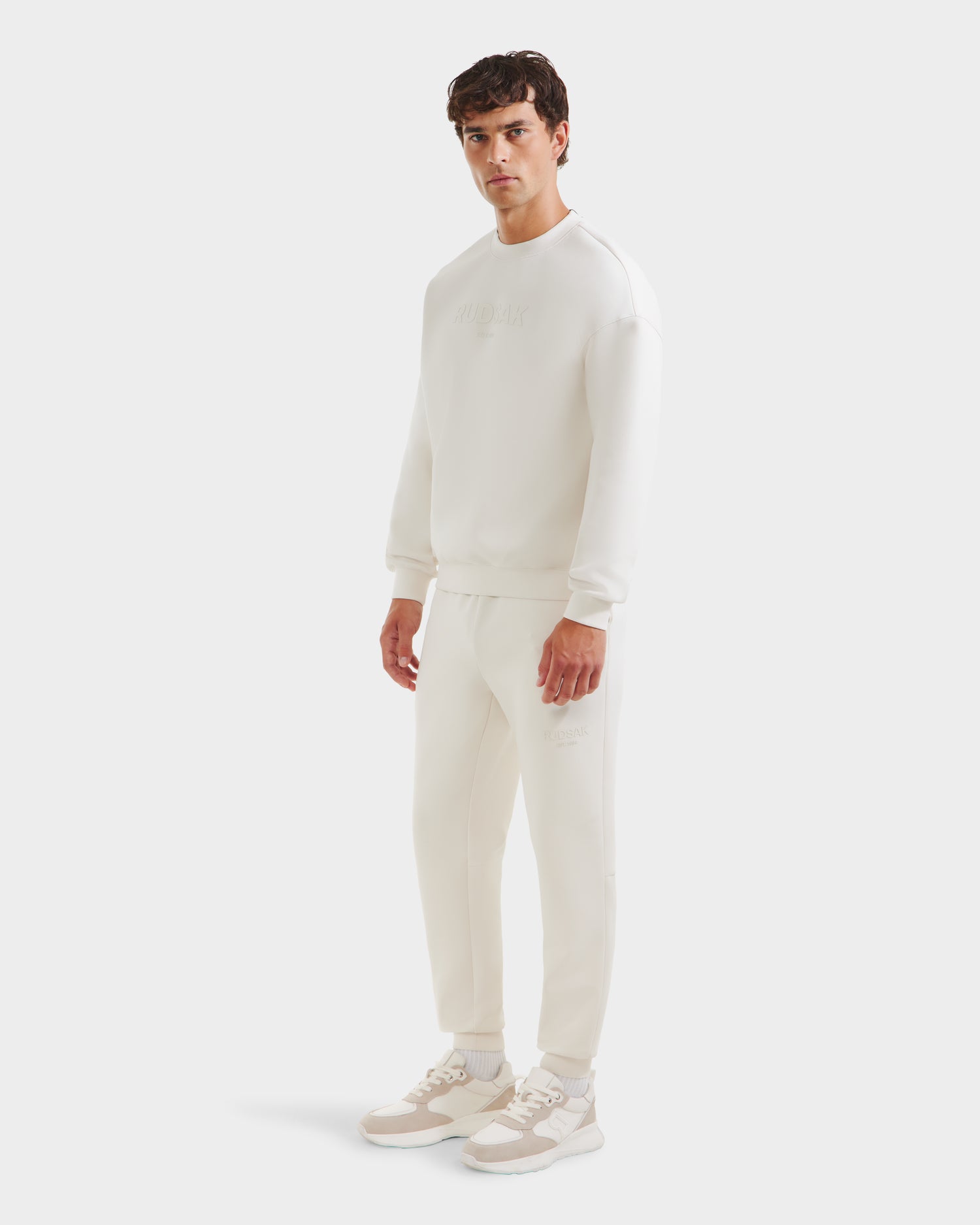 A man stands against a plain white background, wearing the Rudsak BRYN - 9524007 Vanilla M crewneck sweatshirt and jogger pants, paired with white sneakers featuring beige accents.