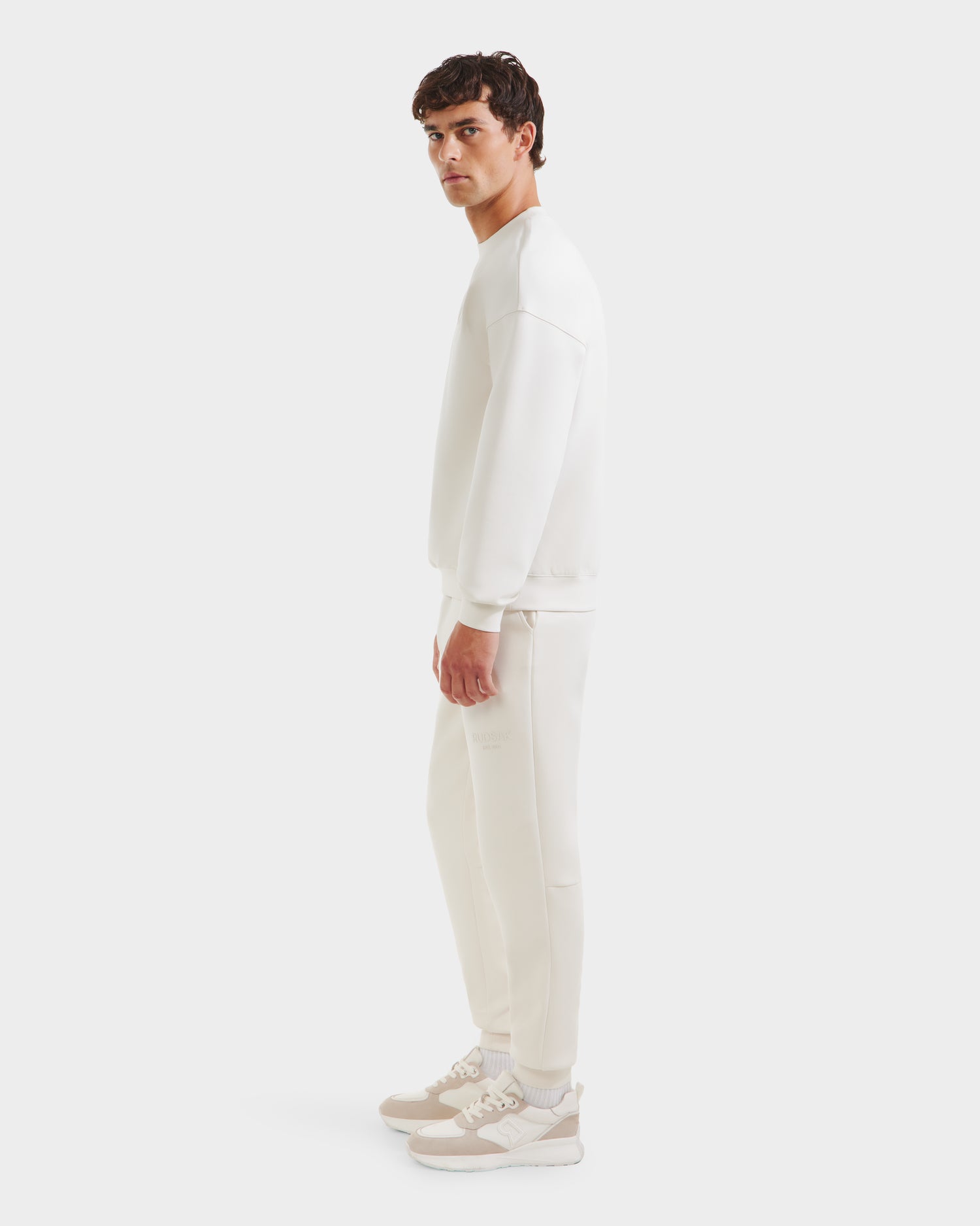 A man in profile against a white background wears the Rudsak BRYN - 9524007 Vanilla M crewneck sweatshirt and jogger pants with beige-and-white sneakers—a genderless, everyday fit.