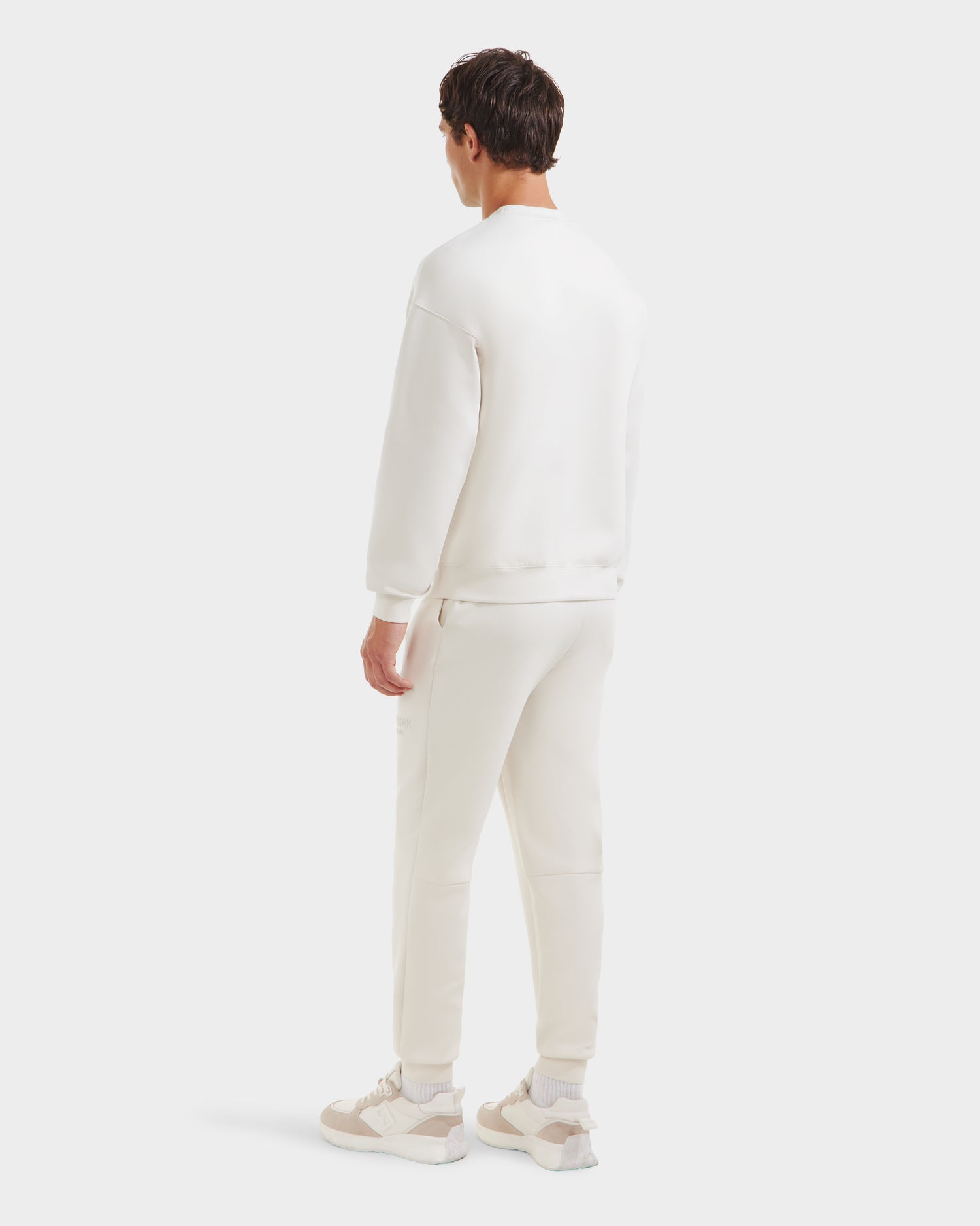 A person faces away from the camera wearing the Rudsak BRYN - 9524007 Vanilla M crewneck sweatshirt and sweatpants, styled with white sneakers featuring beige accents, set against a plain white background.