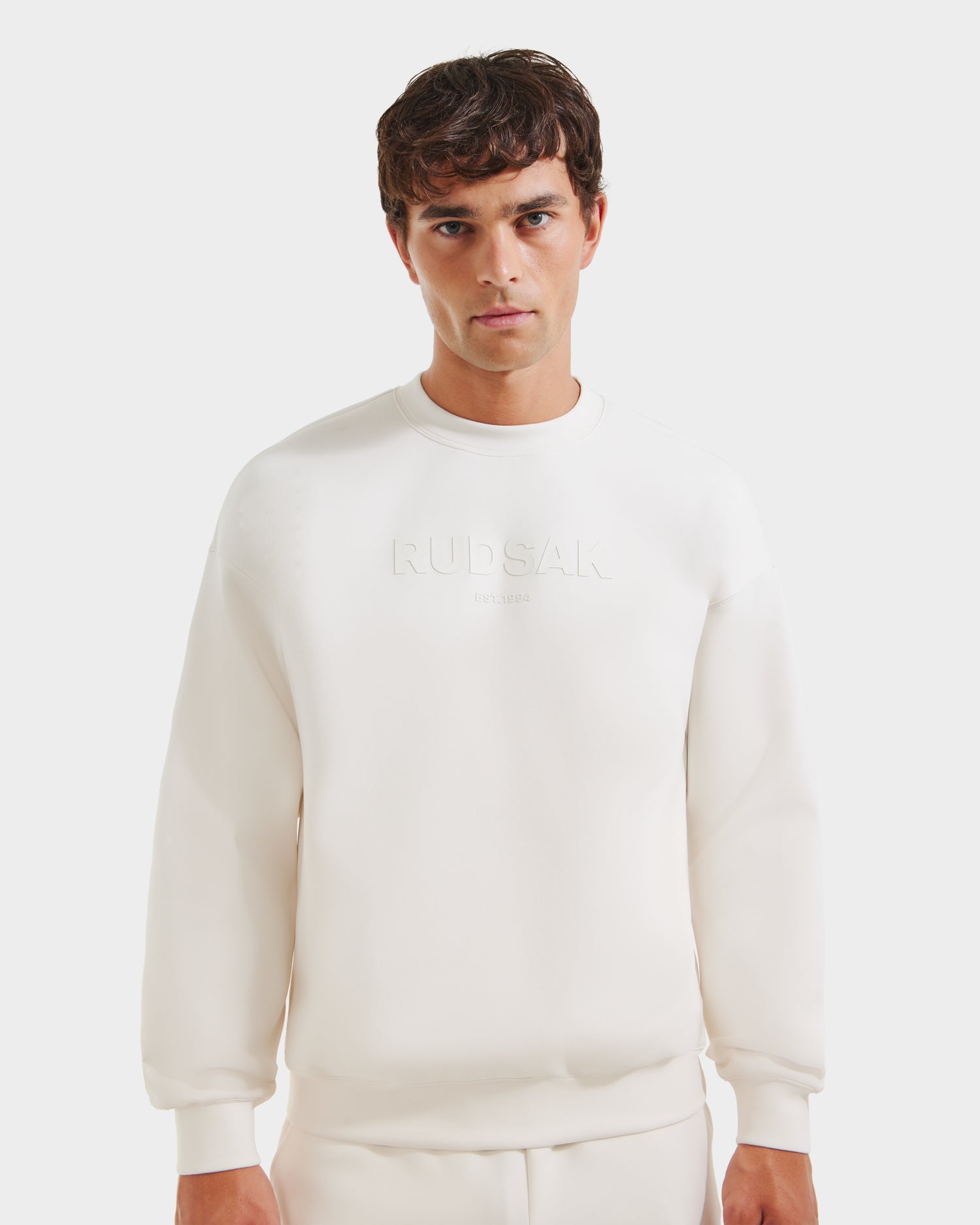 A man wears Rudsak’s BRYN - 9524007 Vanilla M cotton crewneck sweatshirt and pants, featuring embossed RUDSAK on the chest and a relaxed fit, standing against a plain white background.