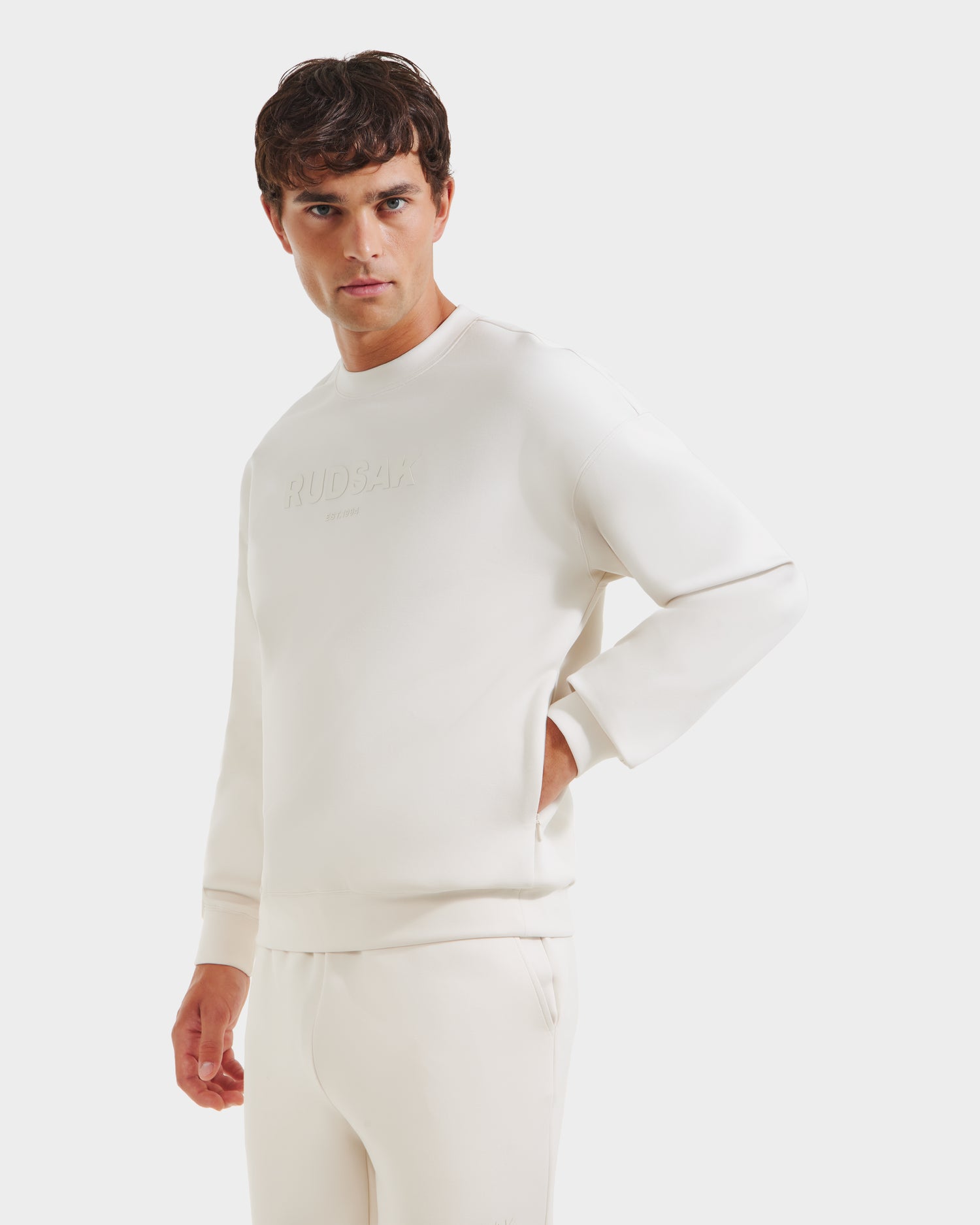 A man models the Rudsak BRYN - 9524007 Vanilla M set—cotton crewneck sweatshirt and matching pants—in front of a white backdrop, gazing left with one hand in his pocket. Embossed text and genderless sizing ensure an effortless fit.