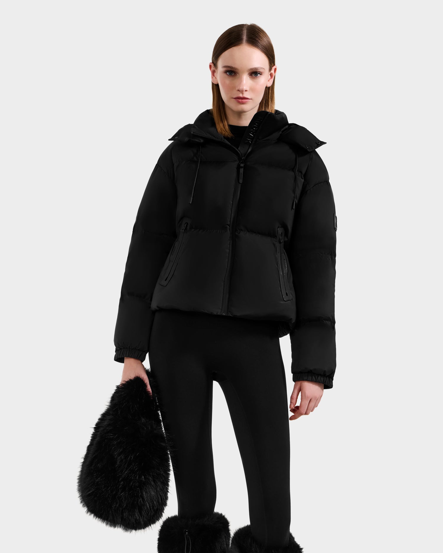 A woman in a black puffer jacket, leggings, and furry boots carries the Rudsak CABAS - 8325545 Black faux fur mini tote, making a statement against a plain light gray background.