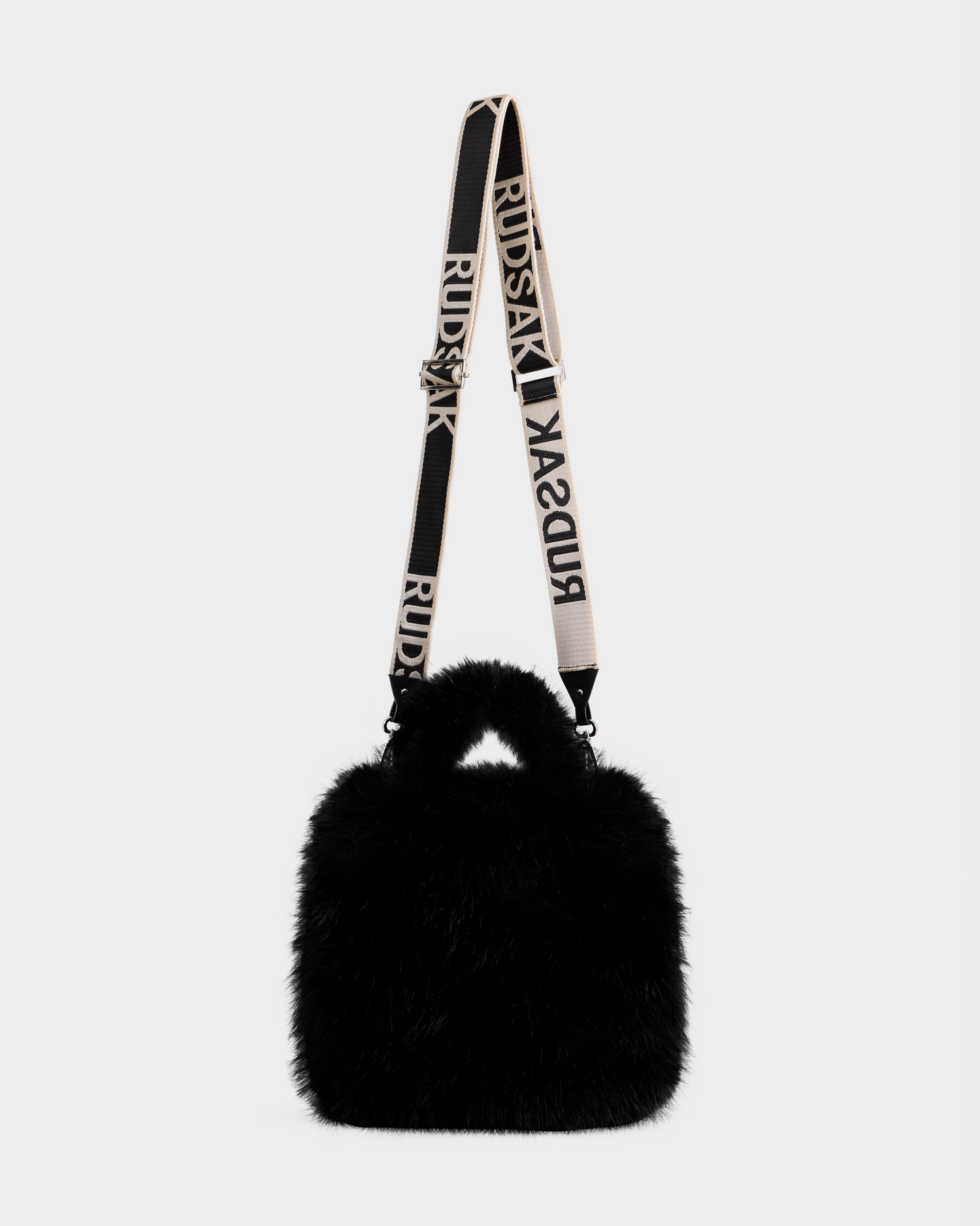 The CABAS - 8325545 Black by Rudsak is a black, fluffy faux fur mini tote with short handles and a wide detachable strap boldly reading RUDSAK—an eye-catching accessory to elevate winter looks, shown on a plain light grey background.