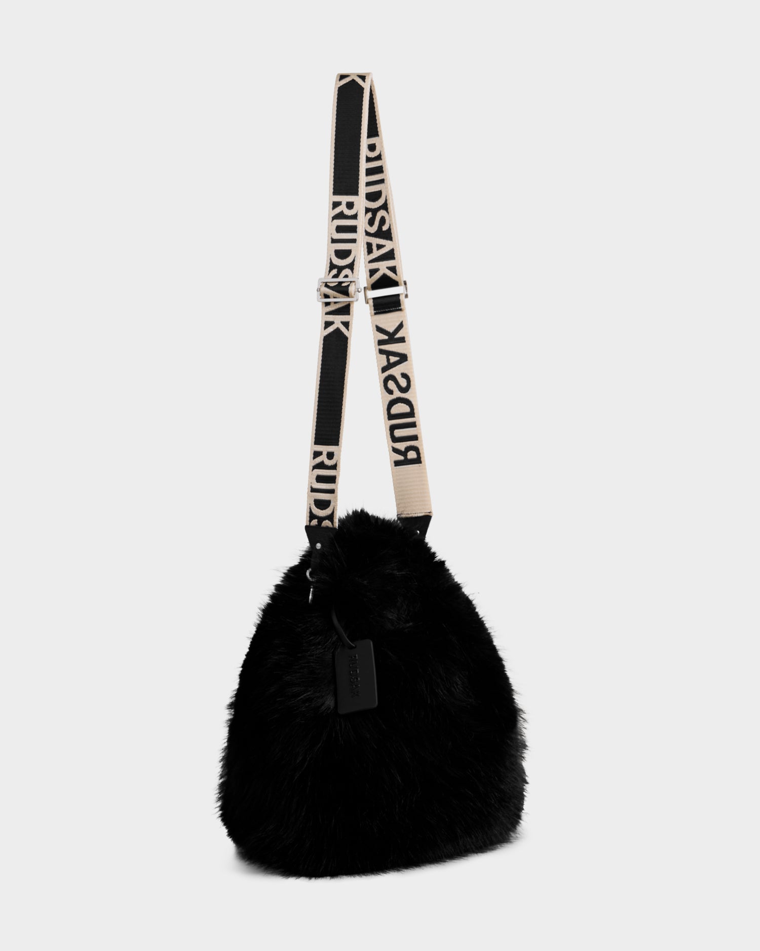 The Rudsak CABAS - 8325545 Black is a black faux fur mini tote with a wide, adjustable cream and black strap featuring bold RUDSAK lettering, and a rectangular black tag on the handle—a soft, plush statement accessory.