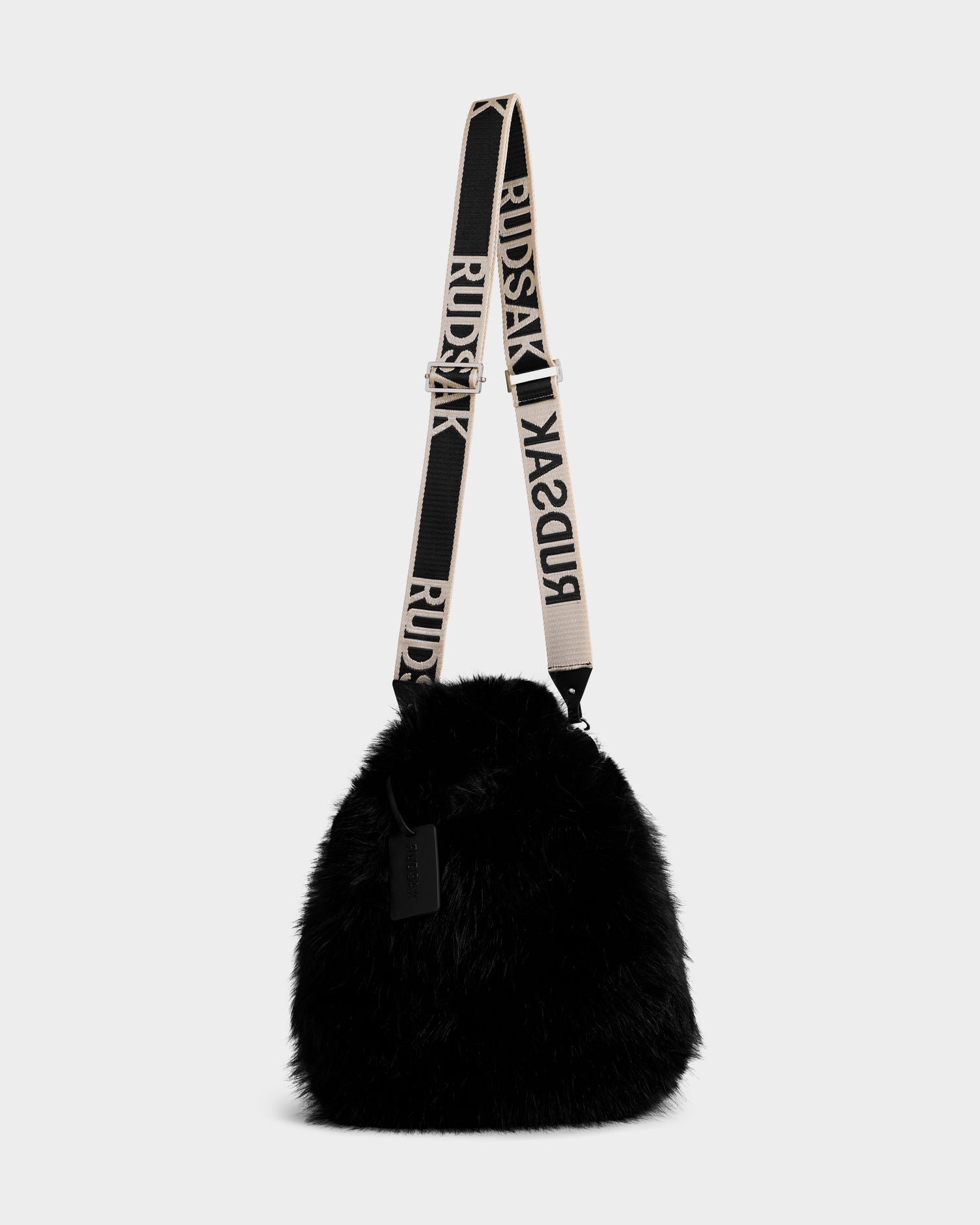 The Rudsak CABAS - 8325545 Black is a black faux fur mini tote featuring a wide adjustable beige strap with bold black RUDSAK text, a soft fluffy texture, and a black tag—perfect as a statement winter accessory.