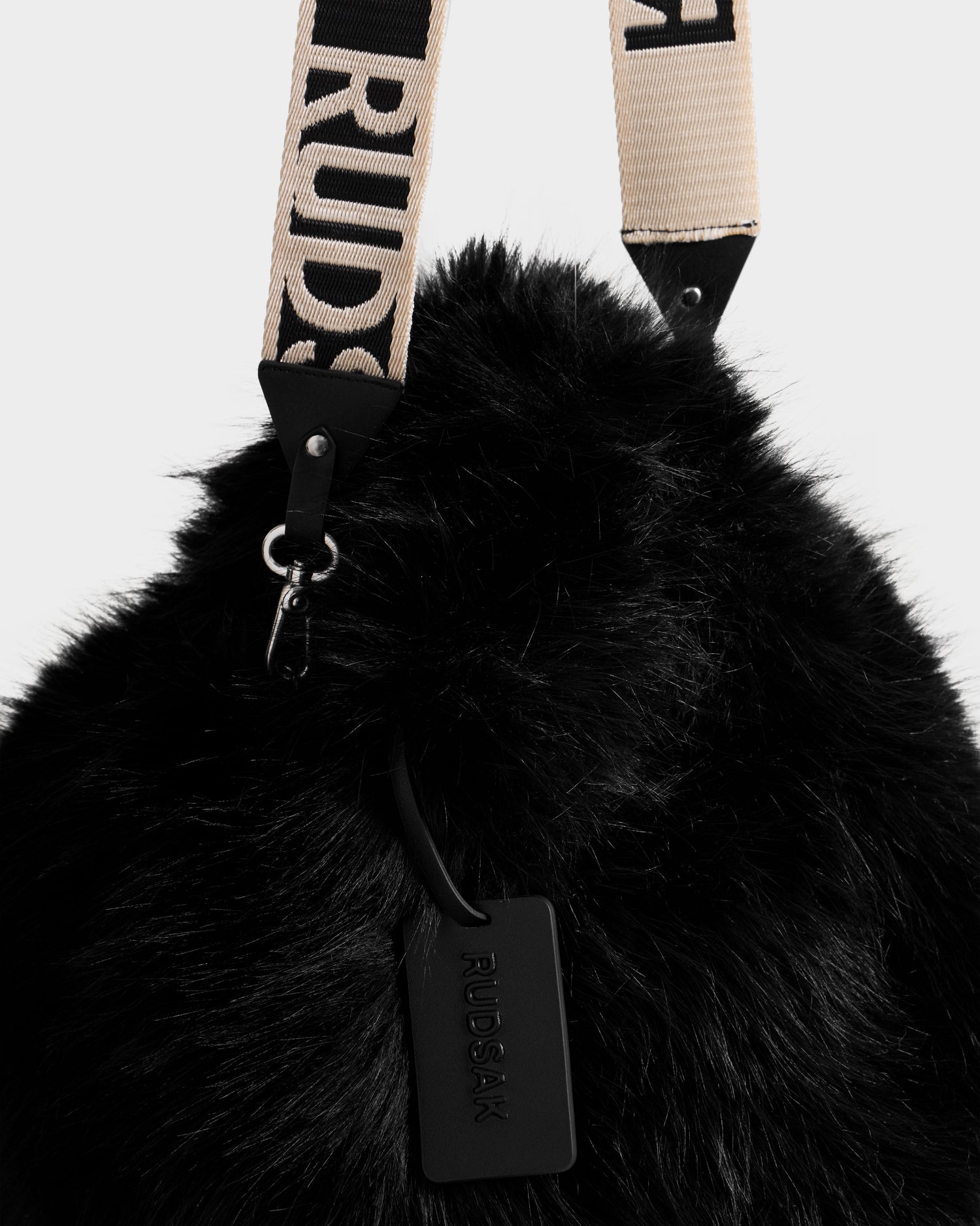 Close-up of the CABAS - 8325545 Black mini tote by Rudsak, featuring black faux fur, a beige and black RUDSAK-branded strap, and a black embossed tag—a chic statement accessory for winter layers.