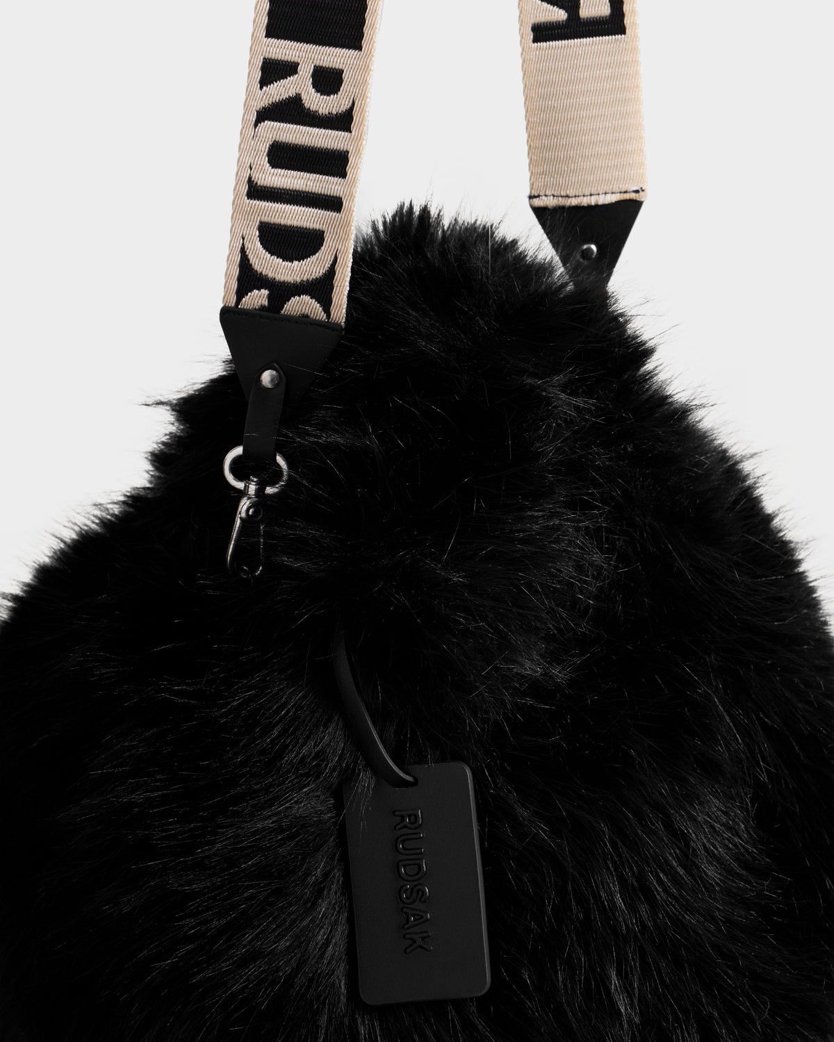 Close-up of the CABAS - 8325545 Black mini tote by Rudsak, featuring black faux fur, a beige and black RUDSAK-branded strap, and a black embossed tag—a chic statement accessory for winter layers.