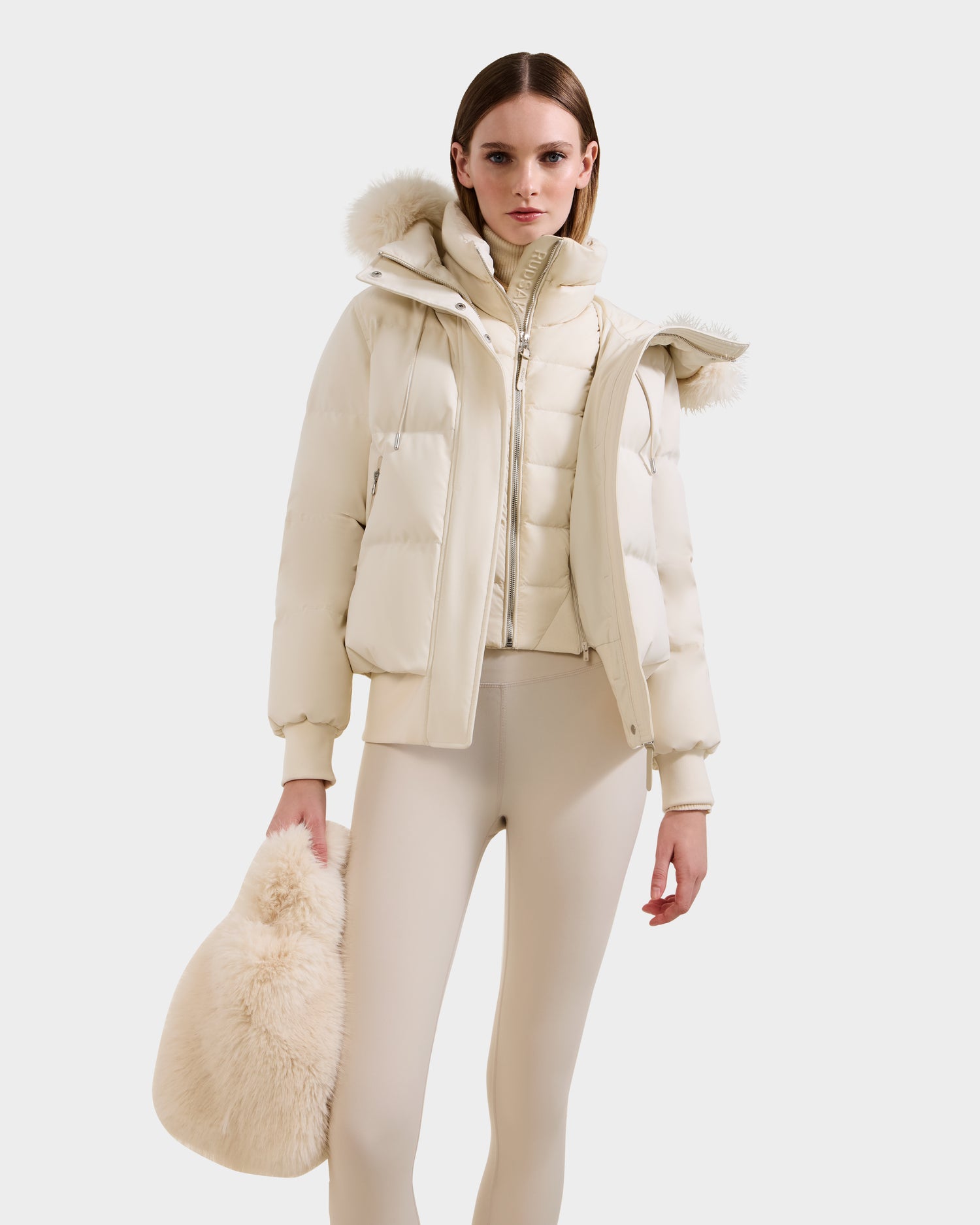 A woman holding the Rudsak CABAS - 8325545 Vanilla plush handbag stands against a white background, dressed in a cream puffer jacket with fur-trimmed hood and matching fitted pants.