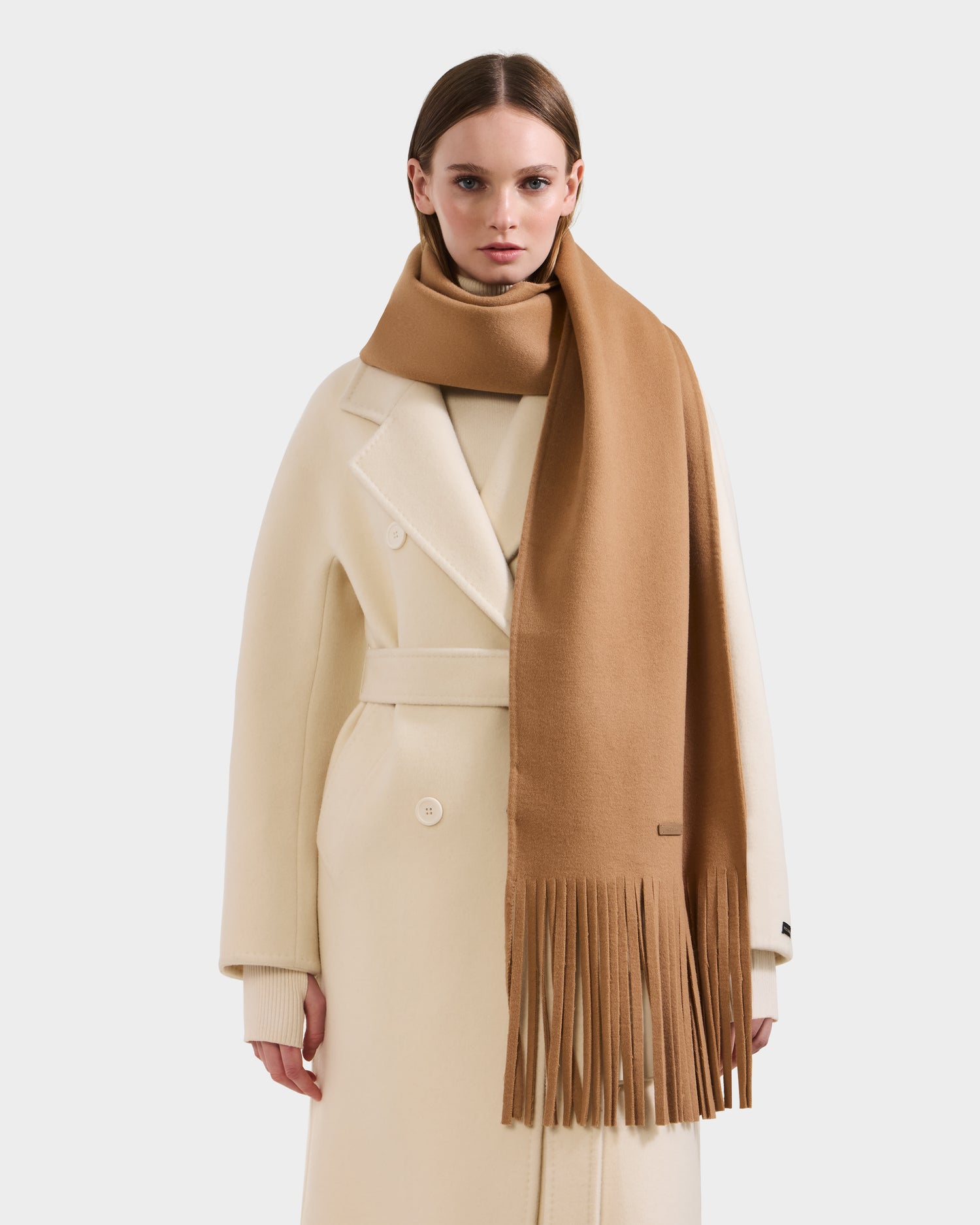A woman in the Rudsak CALEN - 8425532 Camel long belted coat and a Rudsak unisex wool blend scarf with long fringe stands against a white background—true cold-weather essentials.