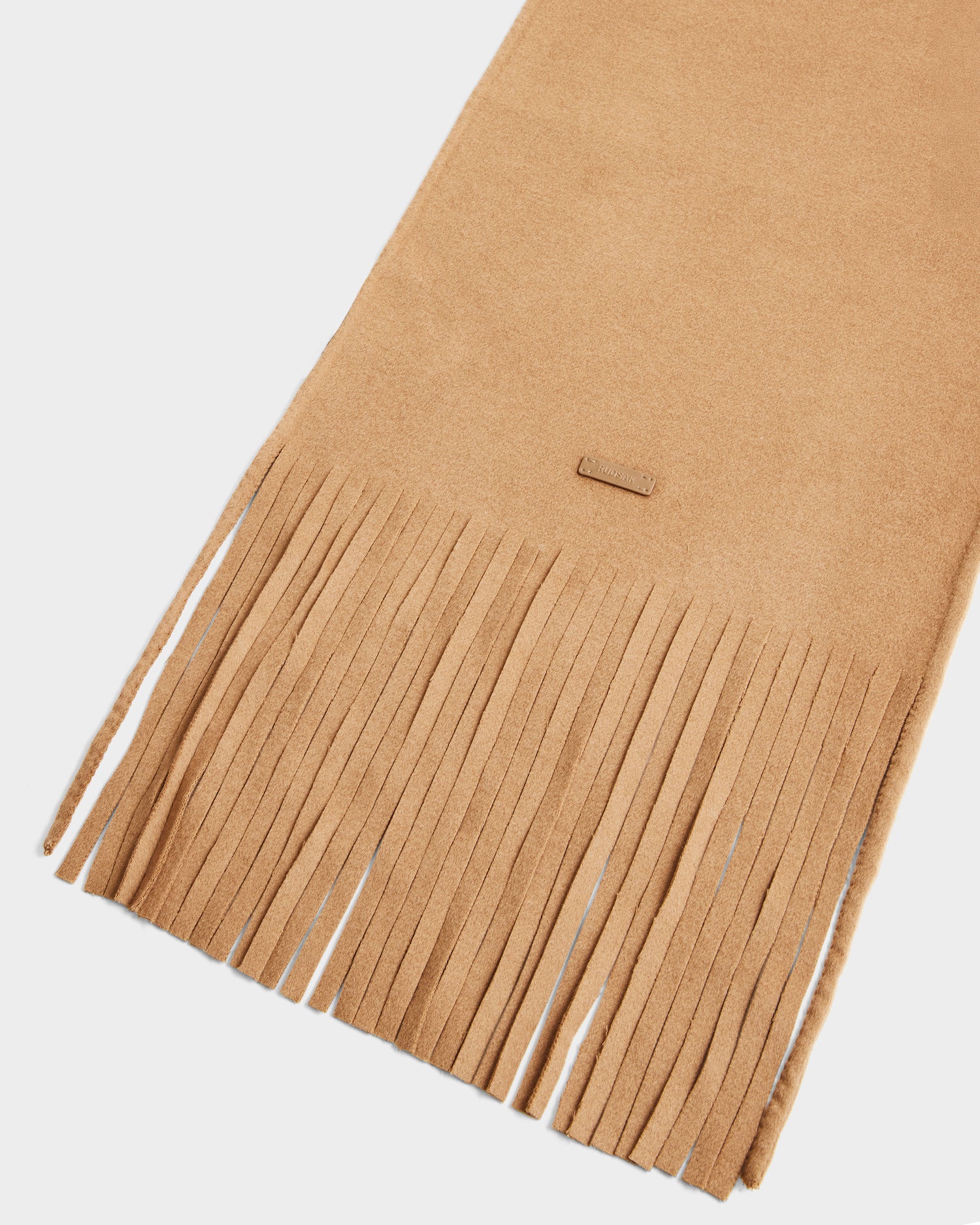 A close-up of the Rudsak CALEN - 8425532 Camel scarf, featuring tan wool blend fabric, long even fringe, and a small rectangular stitched label near the edge.