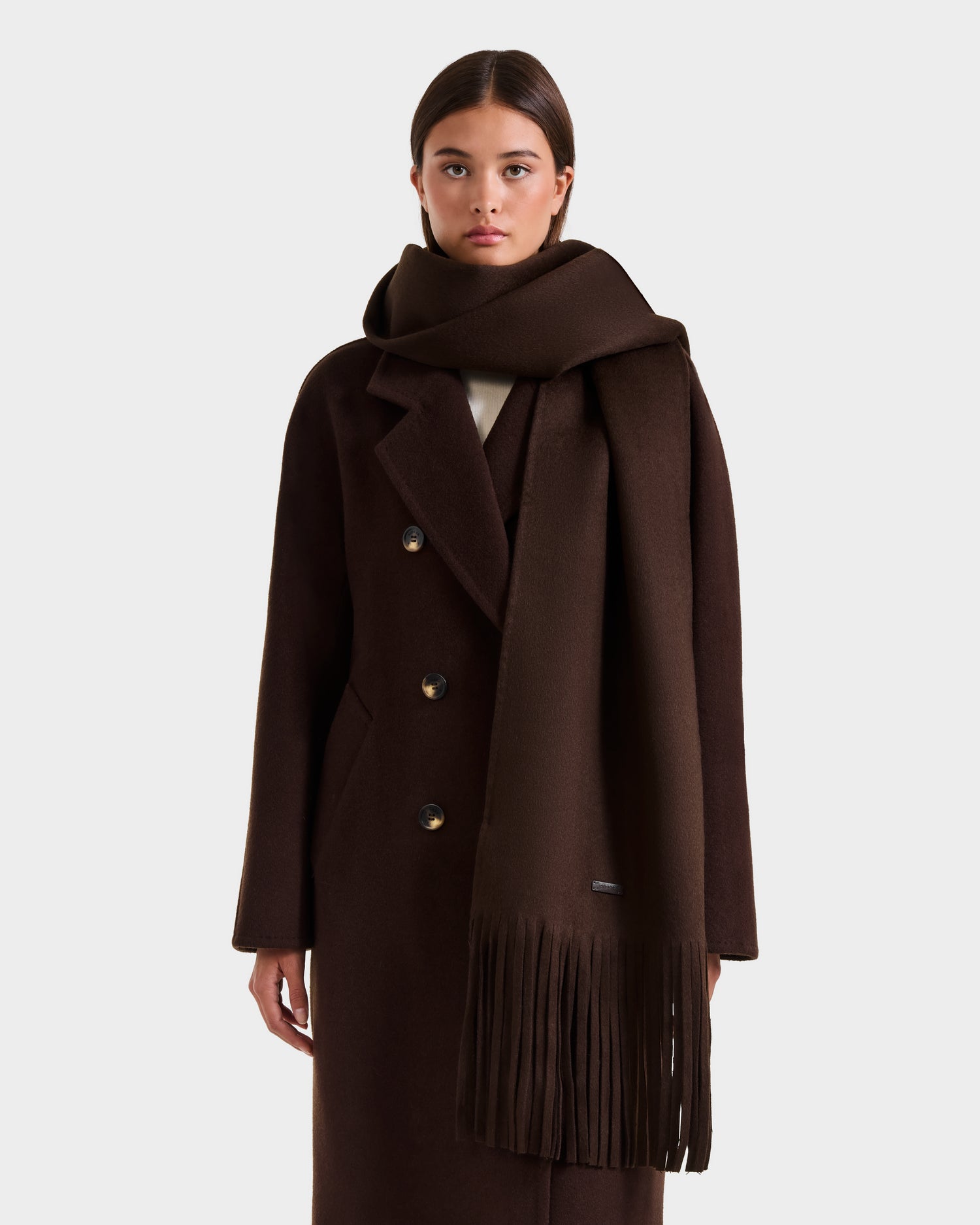 A woman stands against a plain white background wearing a long, dark brown CALEN - 8425532 Espresso coat and a matching Rudsak unisex wool blend scarf with fringed ends—a must-have for cold weather.