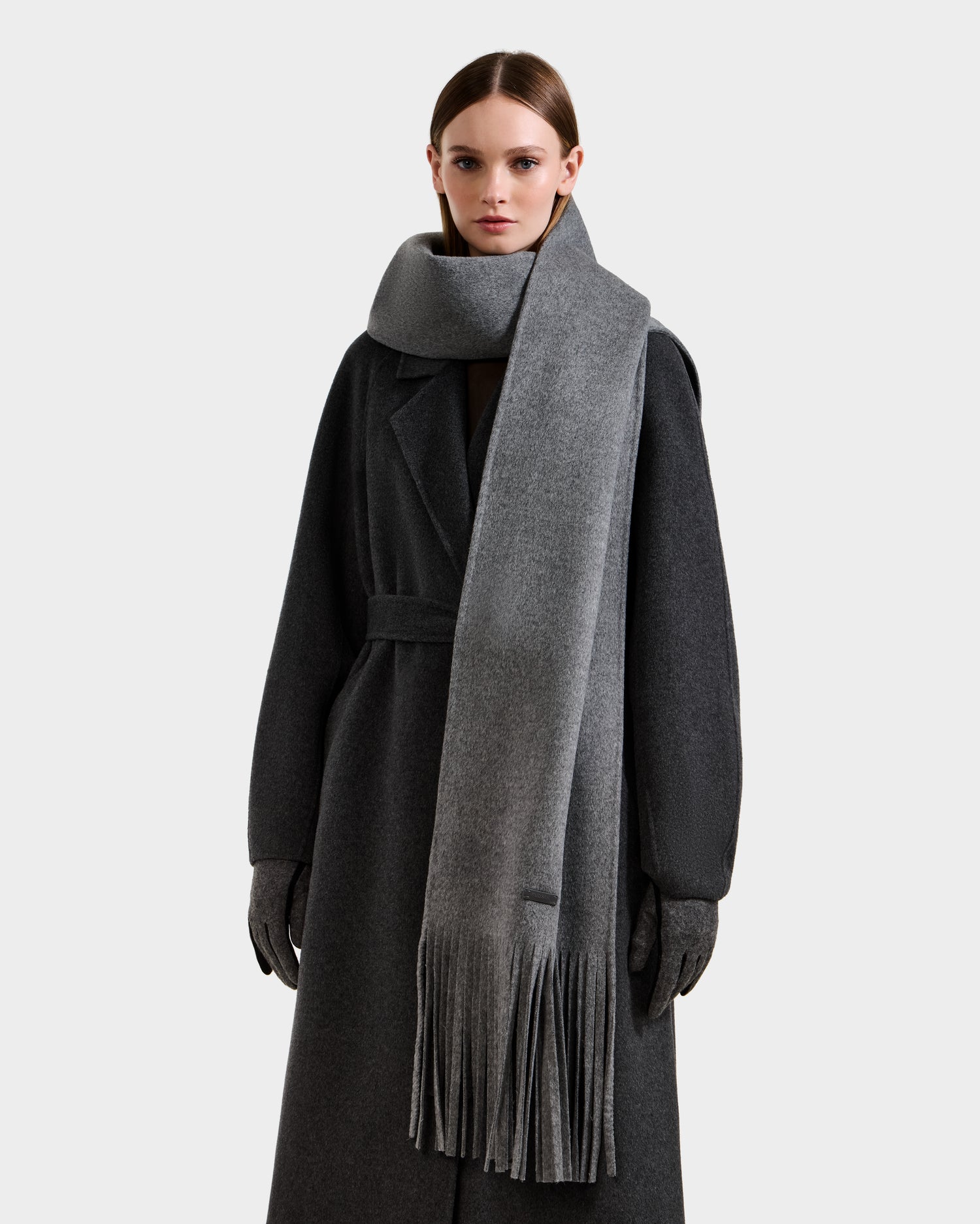 A woman in a long, dark grey coat and gloves stands against a light background, wearing the Rudsak CALEN - 8425532 Grey unisex wool scarf with fringe—a stylish and versatile cold-weather accessory.