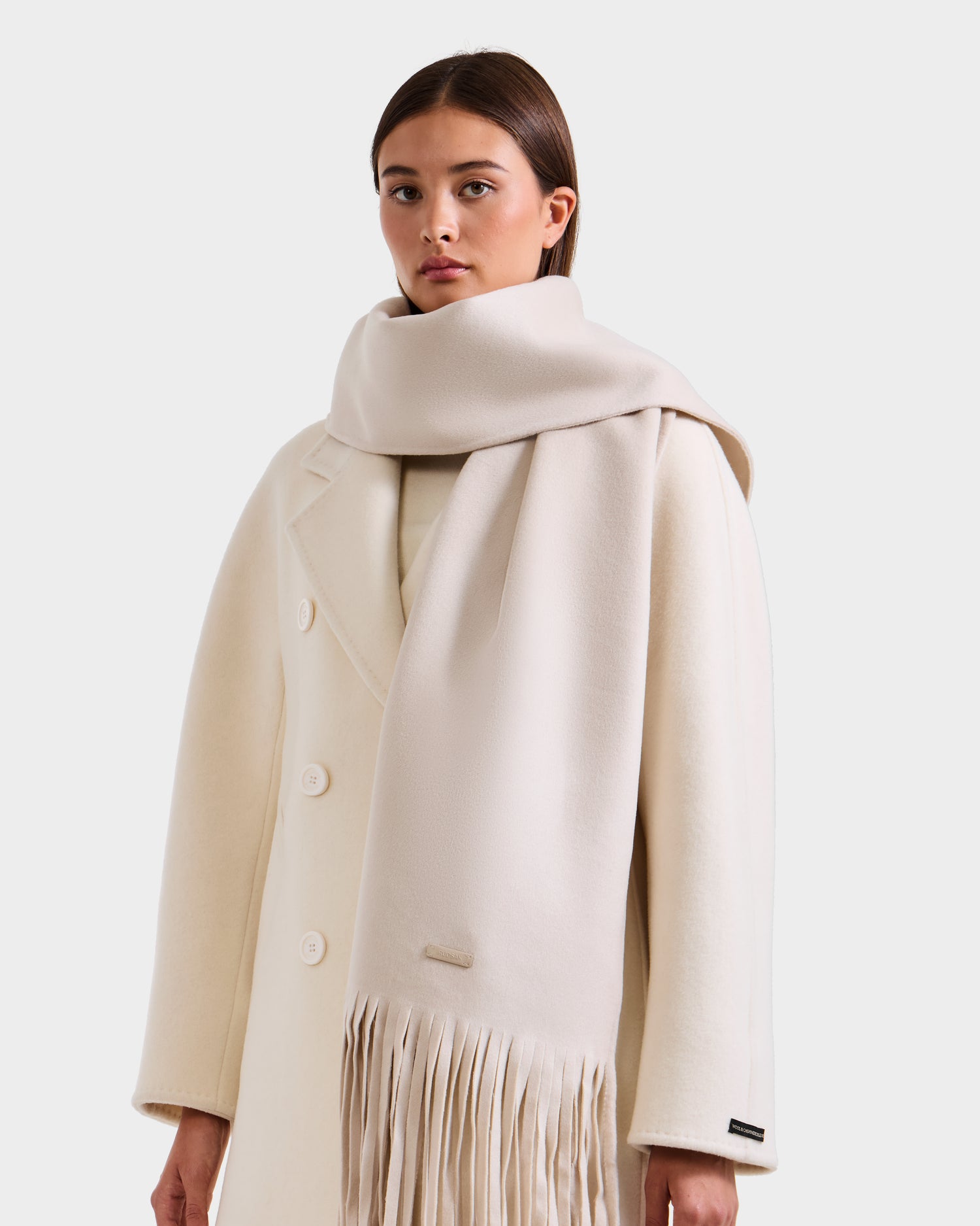 A woman wears the CALEN - 8425532 Vanilla coat by Rudsak with large buttons and a matching fringed wool blend scarf, perfect for cold weather, standing against a plain light background.