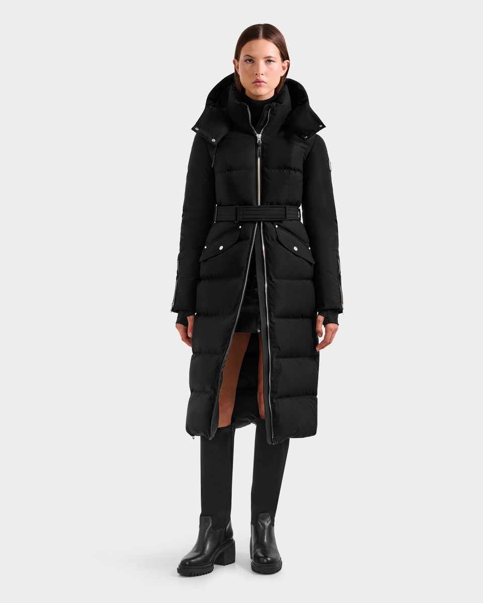 A woman faces forward wearing the Rudsak CALI H - 8125614 Black women's long down parka with a belt and removable hood, styled with a black turtleneck, pants, and boots, set against a plain light gray background.
