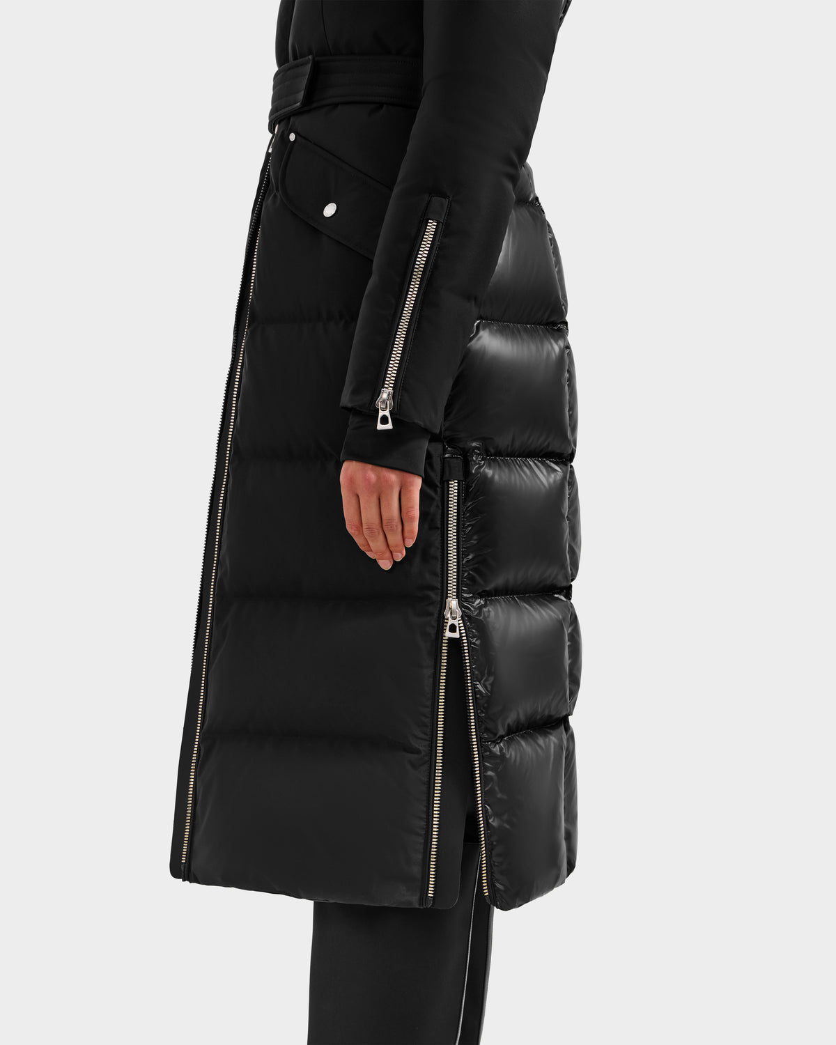 A person wearing the Rudsak CALI H - 8125614 Black women’s long down parka with silver zippers on the sleeves and side. Only the lower body and right arm are visible, showcasing its high-loft 800+ fill power down against a plain light background.