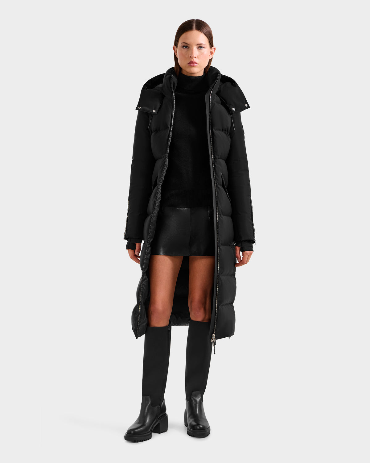 A woman stands facing forward wearing the Rudsak CALI H - 8125614 Black women's long down parka, paired with a black turtleneck, mini skirt, and knee-high boots against a plain light background.