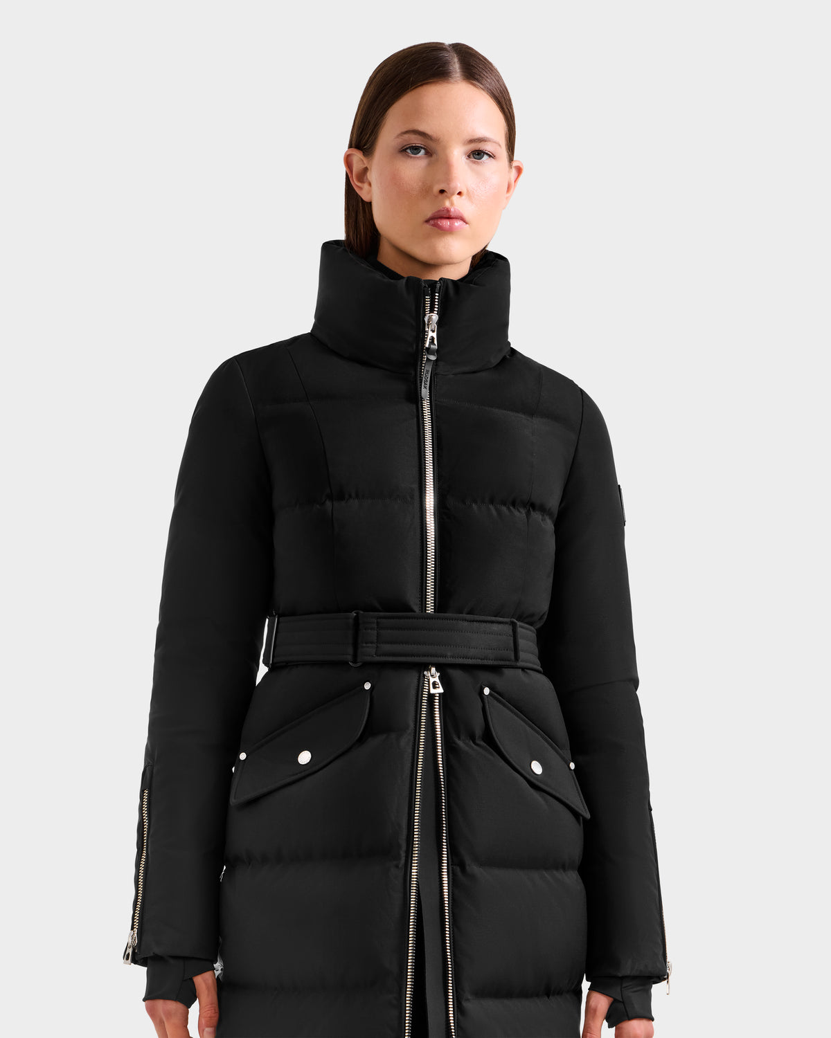 A woman with straight brown hair and a neutral expression wears the Rudsak CALI H - 8125614 Black women’s long down parka, featuring a high collar, silver zippers, and 800+ fill power down, standing against a plain light gray background.