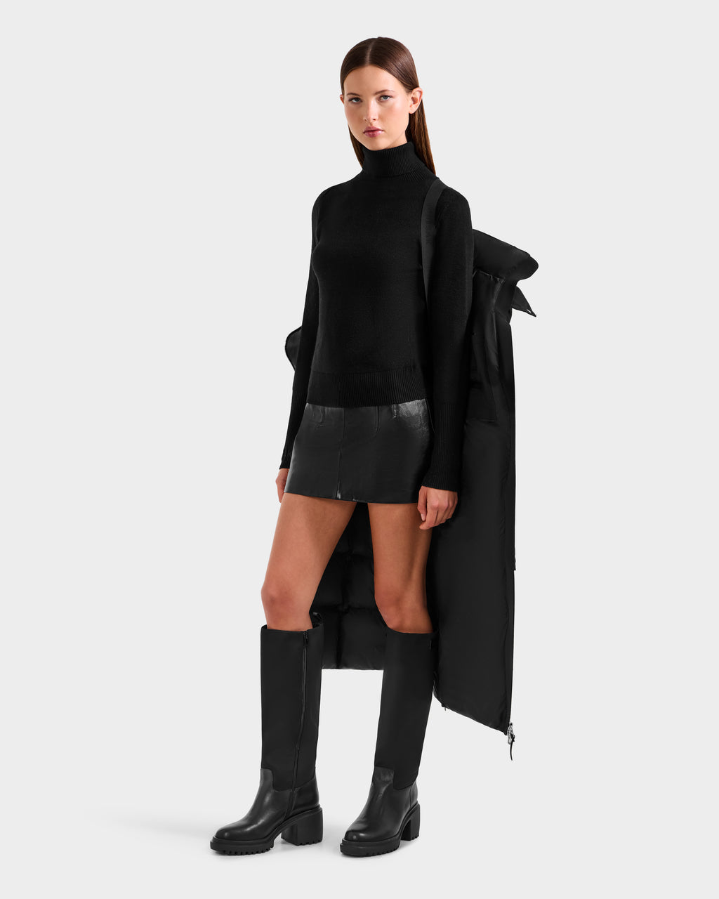 A woman poses against a white background wearing a black turtleneck, short black leather skirt, tall black boots, and the Rudsak CALI H - 8125614 Black women’s long down parka with high-loft 800+ fill power draped over her shoulders.
