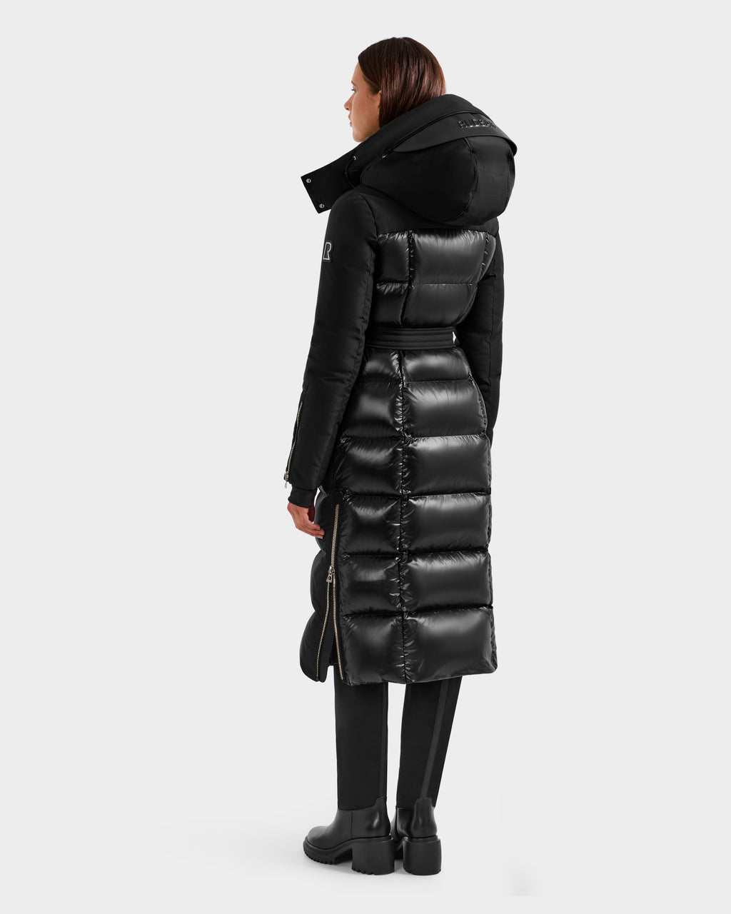 A person faces away in the Rudsak CALI H - 8125614 Black women's long down parka, featuring a removable hood, high-loft 800+ fill power down, side zippers, and a glossy lower section. Paired with black pants and boots; plain light gray background.