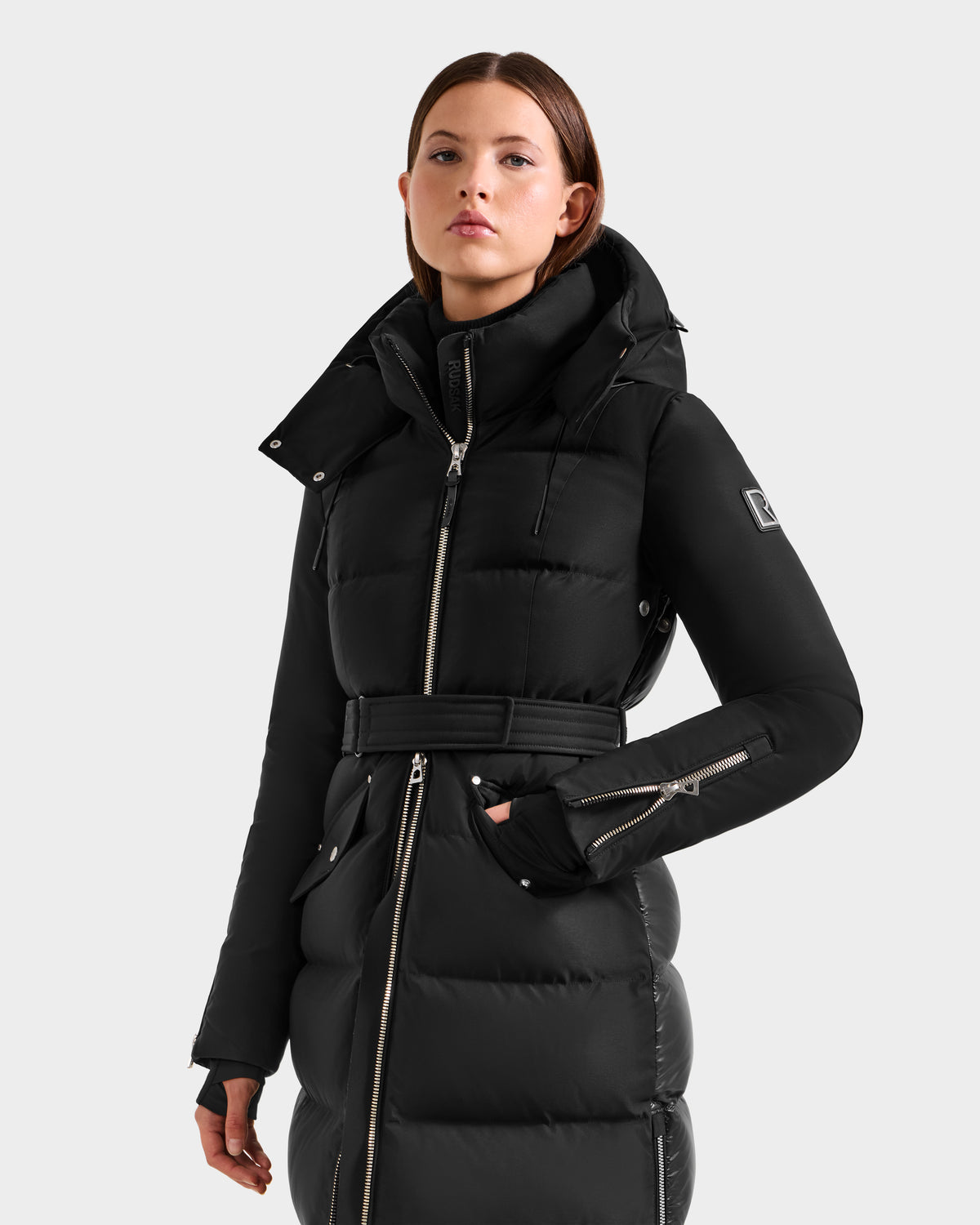 A woman wears the Rudsak CALI H - 8125614 Black fitted long down parka with a high collar, zip detail, removable hood, and belted waist, standing confidently with one hand in her pocket against a plain light background.