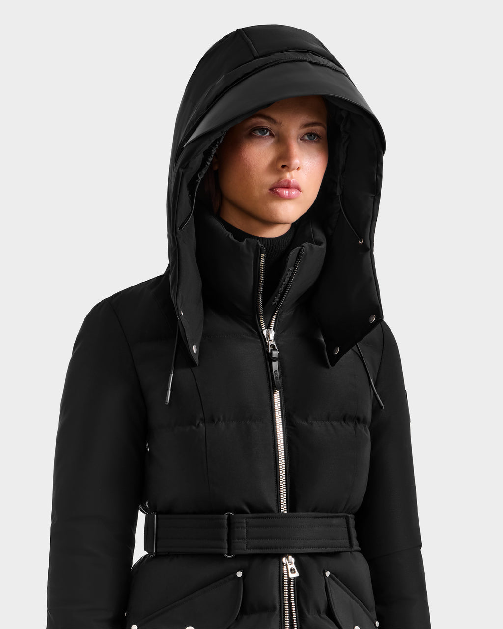 A person stands against a plain light background wearing the Rudsak CALI H - 8125614 Black women’s long down parka featuring a high collar, silver zipper, belted waist, and removable hood.