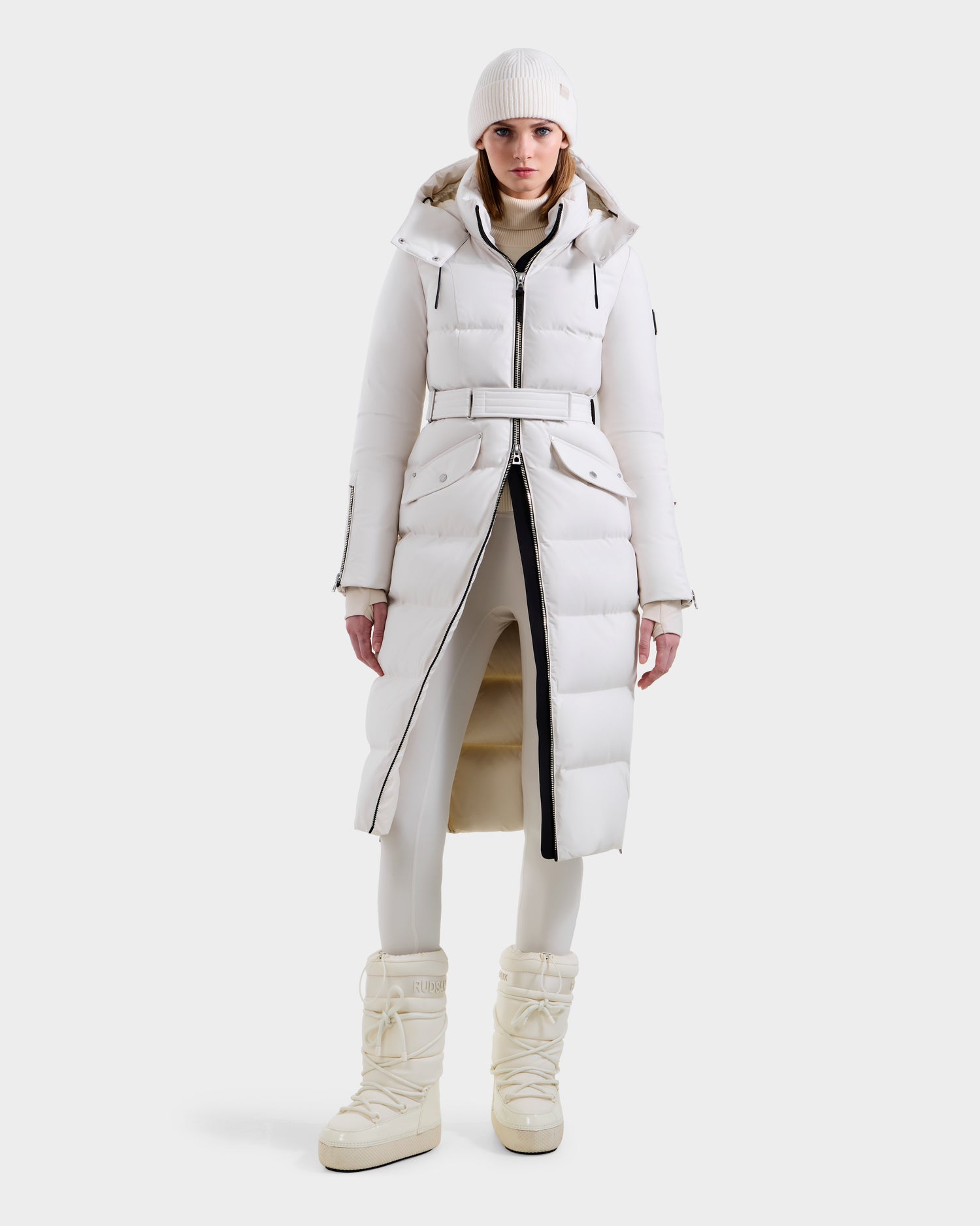 A person faces forward in an all-white winter outfit, featuring the Rudsak CALI H - 8125614 Vanilla women’s long down parka with a removable hood, paired with fitted pants and puffy snow boots, against a plain white background.
