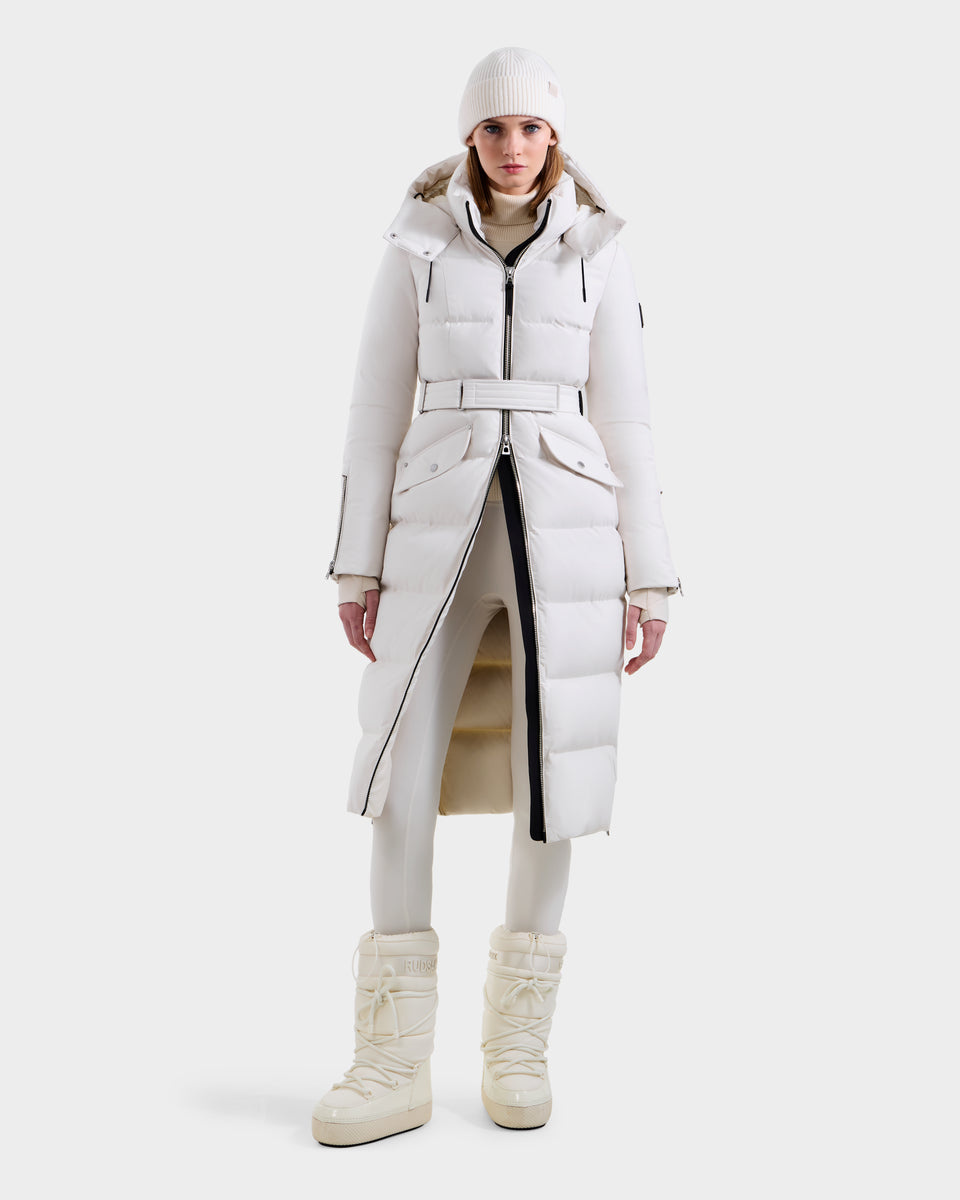 A person faces forward in an all-white winter outfit, featuring the Rudsak CALI H - 8125614 Vanilla women’s long down parka with a removable hood, paired with fitted pants and puffy snow boots, against a plain white background.