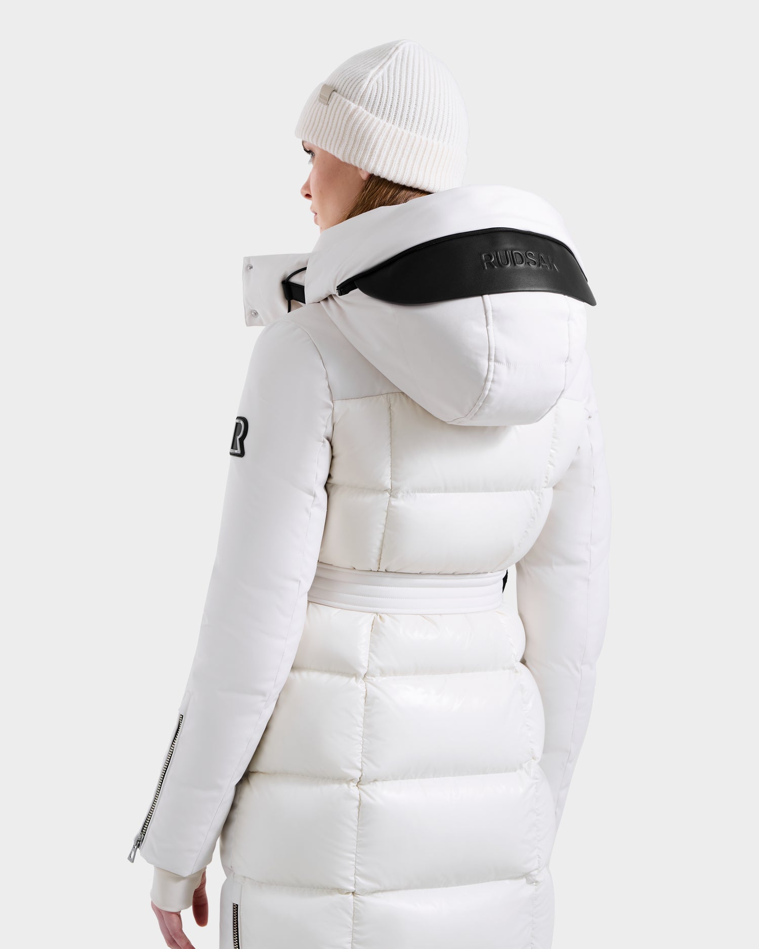 A person in the Rudsak CALI H - 8125614 Vanilla women’s long down parka with a removable hood featuring a black patch and text, and logo on the upper sleeve, wears a white knit beanie, facing away against a plain light gray background.