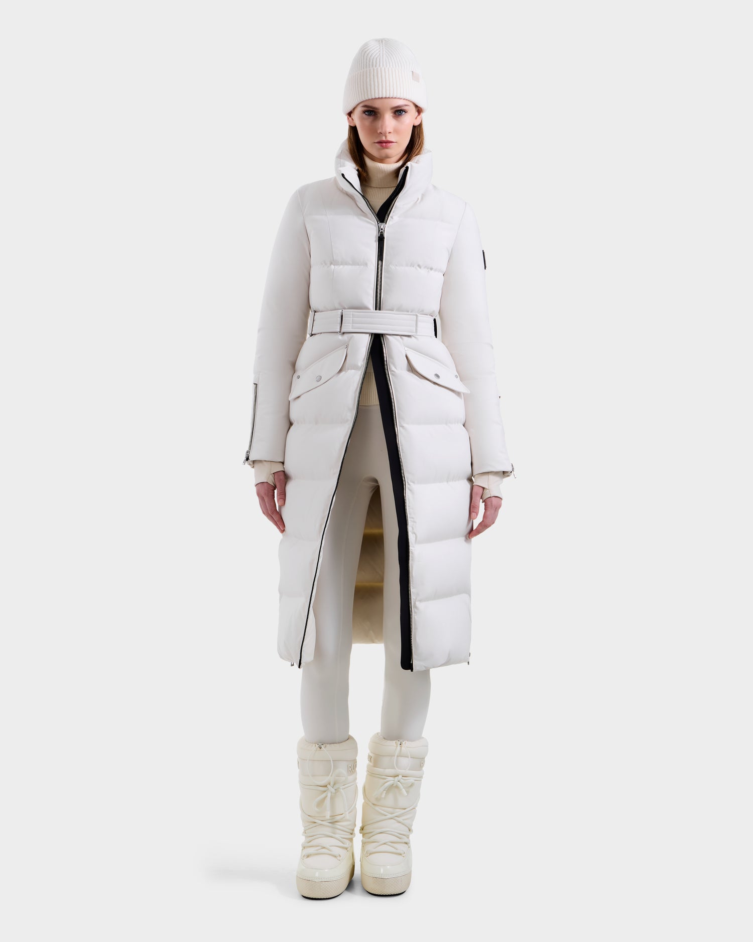 A woman faces forward wearing the Rudsak CALI H - 8125614 Vanilla long down parka with removable hood, styled with white leggings, boots, and a knit beanie for a sleek, modern winter look against a plain white background.