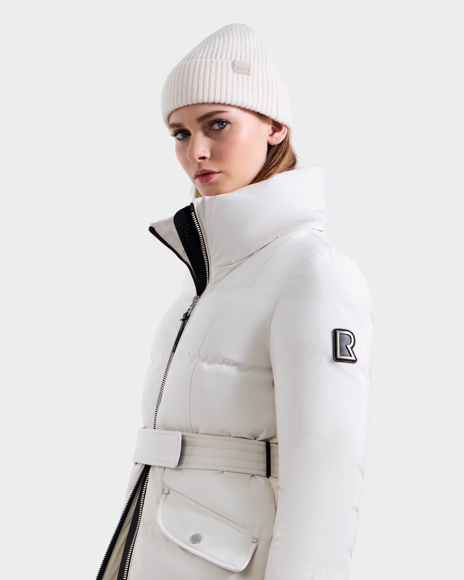 A woman wears the Rudsak CALI H - 8125614 Vanilla women’s long down parka with belt, high collar, and removable hood, plus a white knitted beanie, standing against a plain light gray background, looking at the camera.