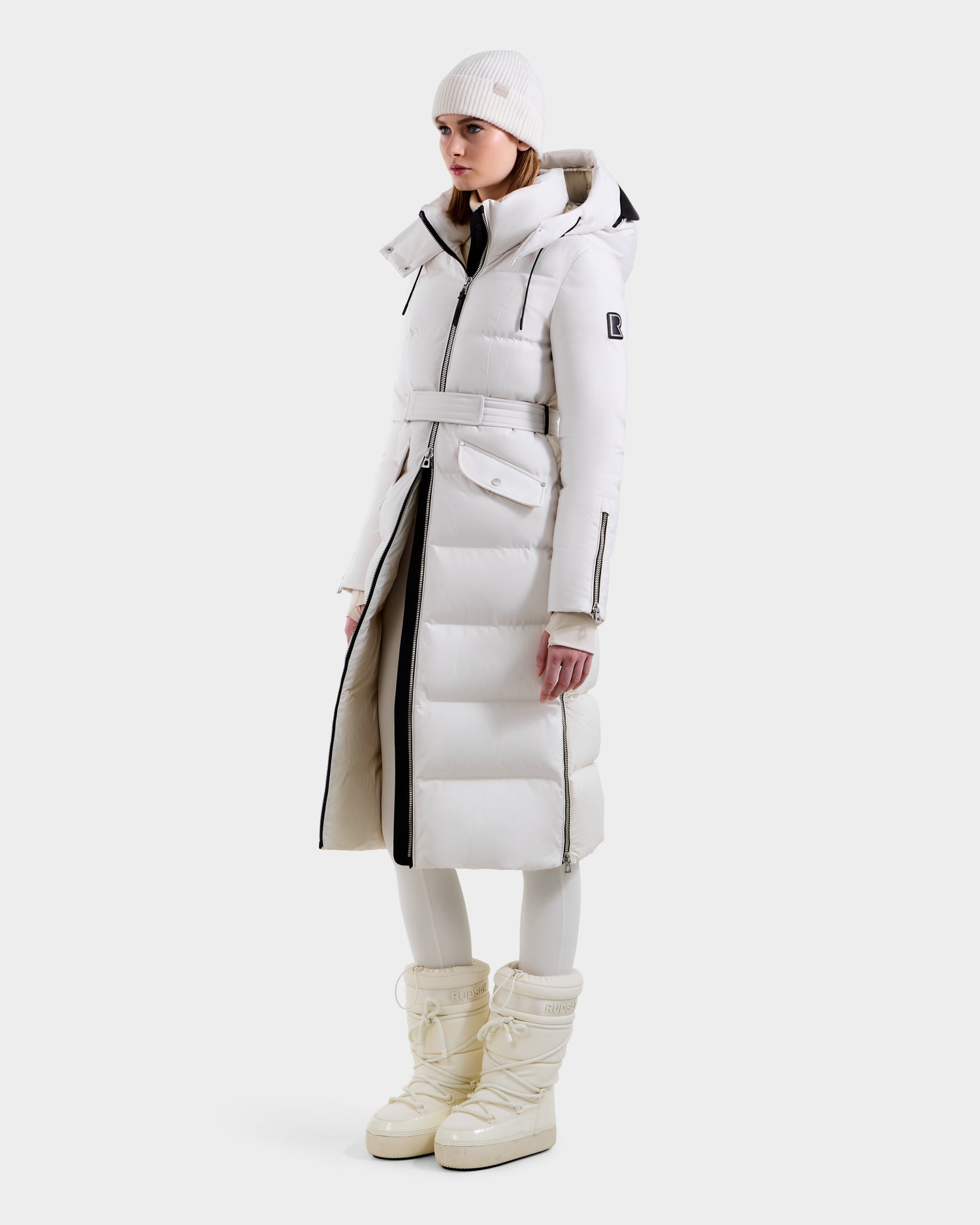 A woman stands against a plain white background wearing the Rudsak CALI H - 8125614 Vanilla long down parka with a belt and removable hood, paired with a white knit beanie, white leggings, and tall white snow boots.