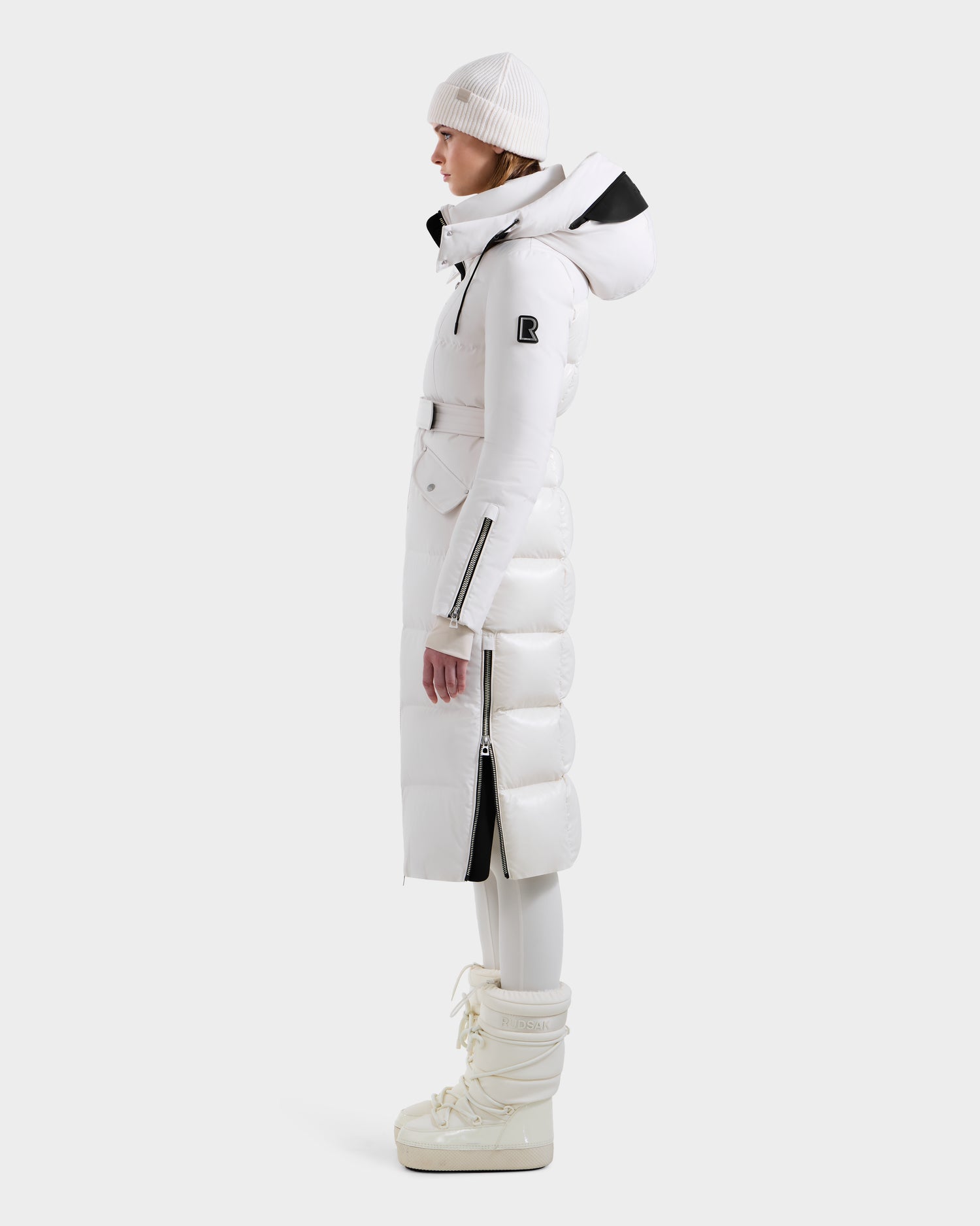 A person in profile models the Rudsak CALI H - 8125614 Vanilla women’s long down parka, accented in black and styled with a white knit hat, white leggings, and white boots, posed against a plain light background.