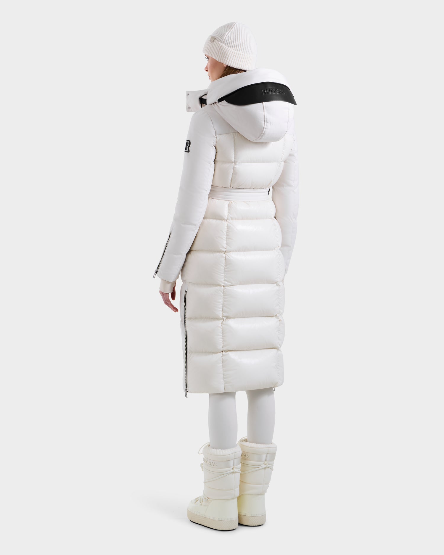 A person stands facing away, wearing Rudsak’s CALI H - 8125614 Vanilla women’s long down parka with a black-accented removable hood, waist belt, white leggings, winter boots, and a white beanie for stylish warmth.
