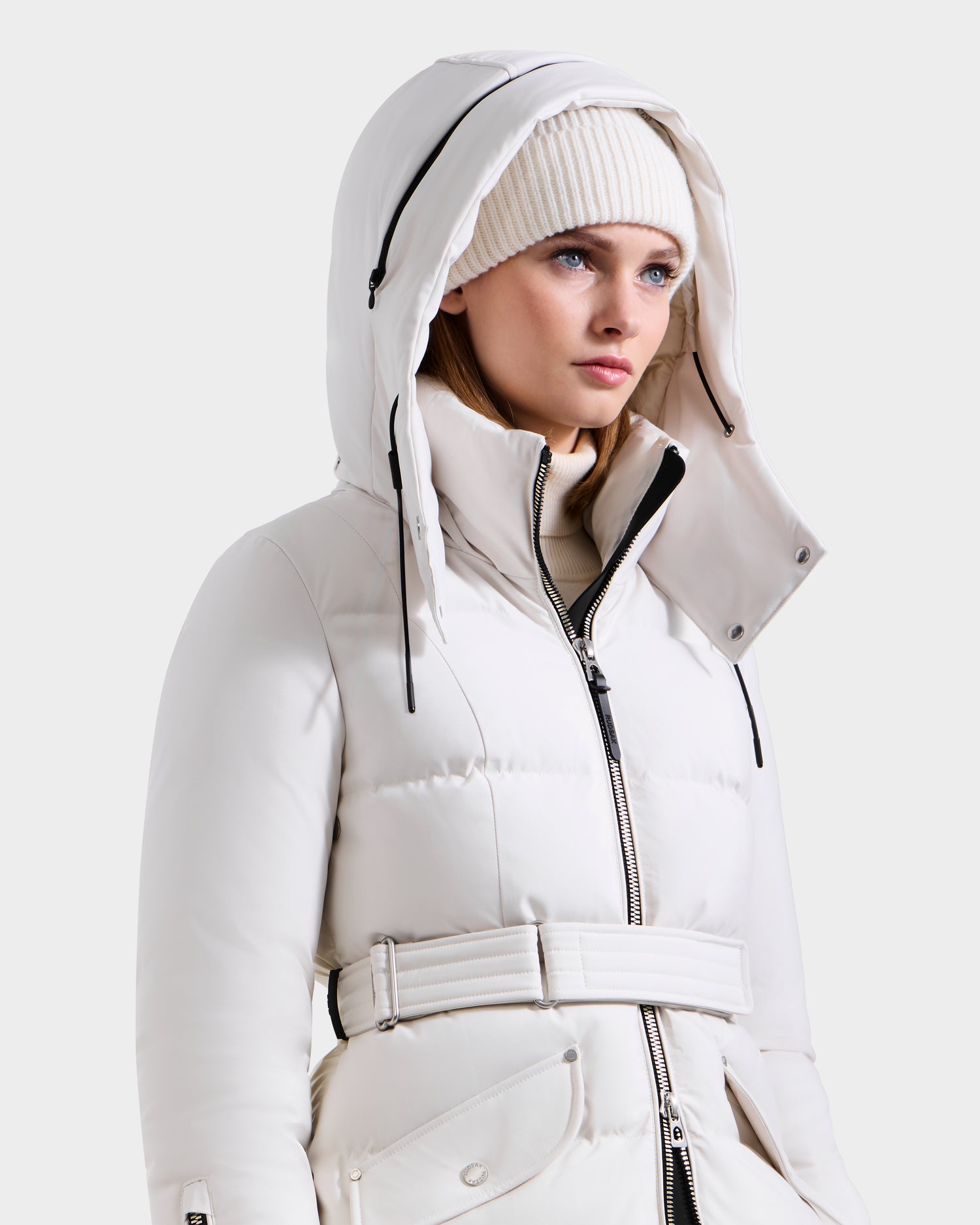 A woman wears the Rudsak CALI H - 8125614 Vanilla long down parka, featuring a belted waist, zippered front, high-loft fill, and large removable hood. She pairs it with a white knit beanie against a plain light gray background.